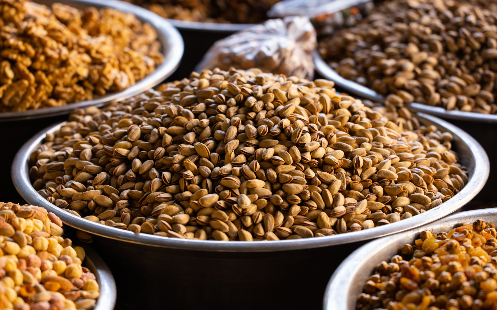 Various roasted nuts and seeds displayed in large metal bowls at a market or store.