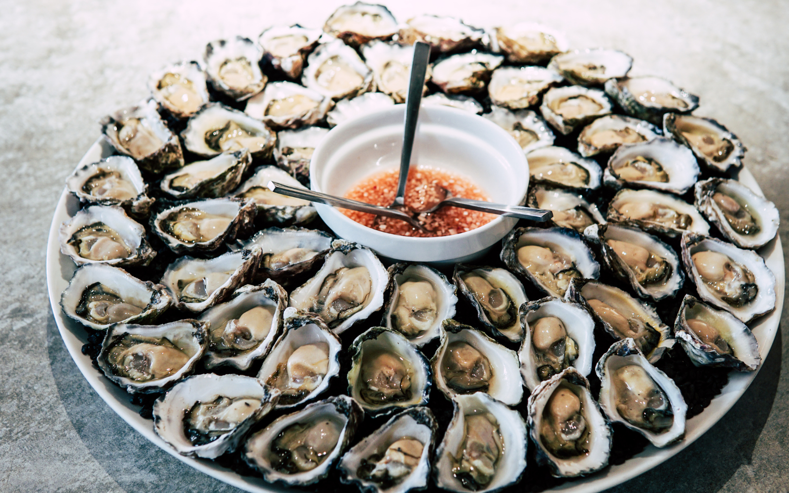 A large round white platter filled with fresh oysters on the half shell, arranged in a circle around a small bowl of cocktail sauce. The platter is on a gray textured surface.