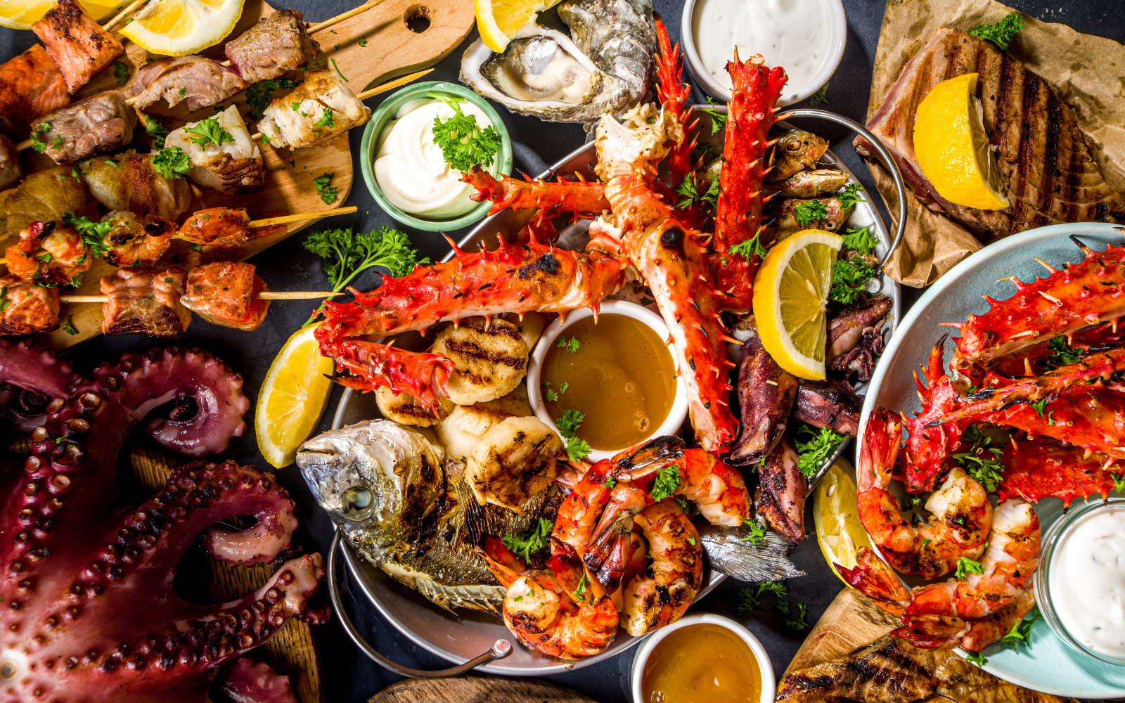 Seafood platter including lobster claws, shrimp, oysters, octopus, grilled fish, and various dipping sauces, garnished with lemon wedges and fresh herbs.