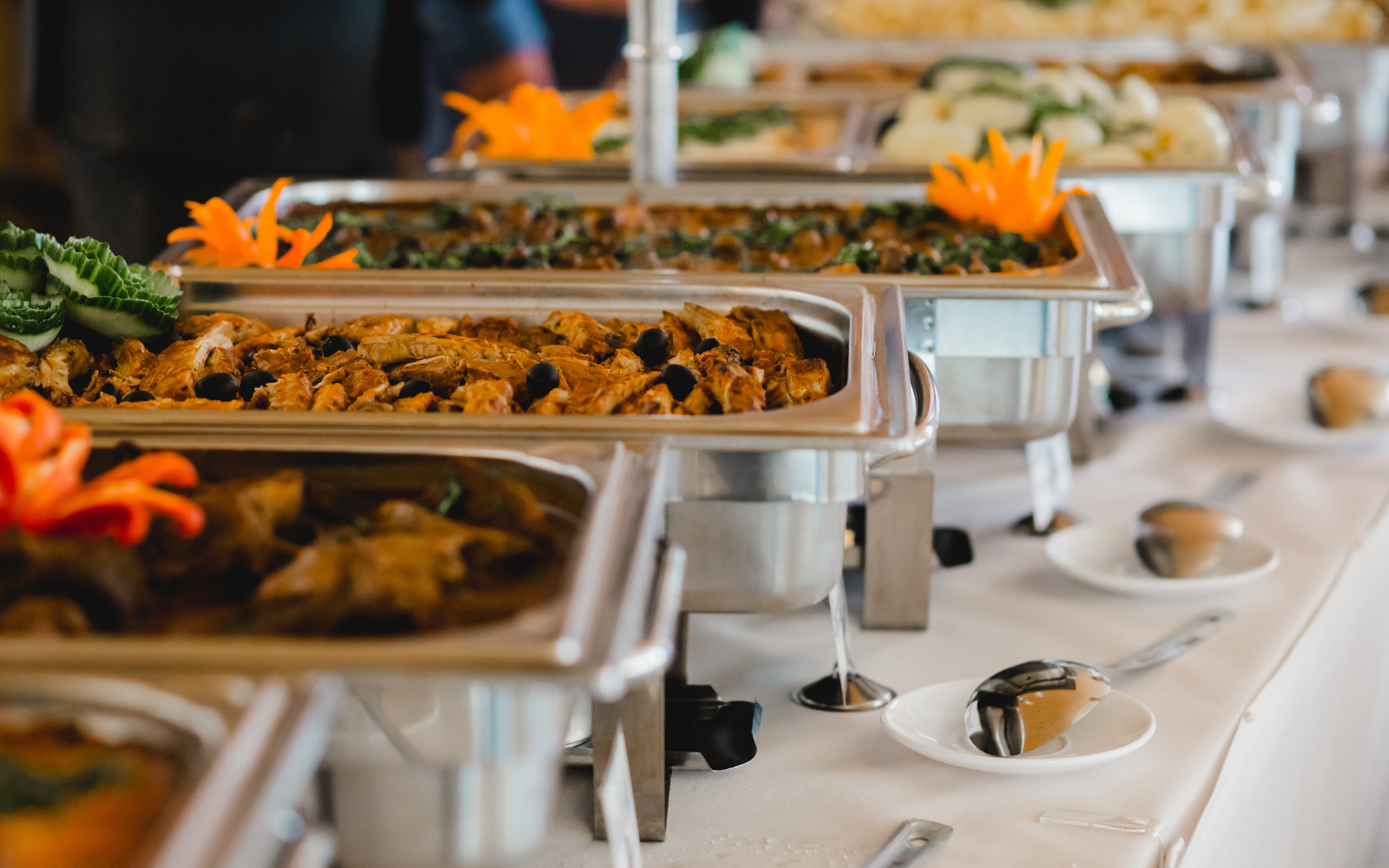 Buffet style food with chafing dishes containing grilled chicken, vegetables, and other dishes on a white tablecloth with serving utensils and plates.