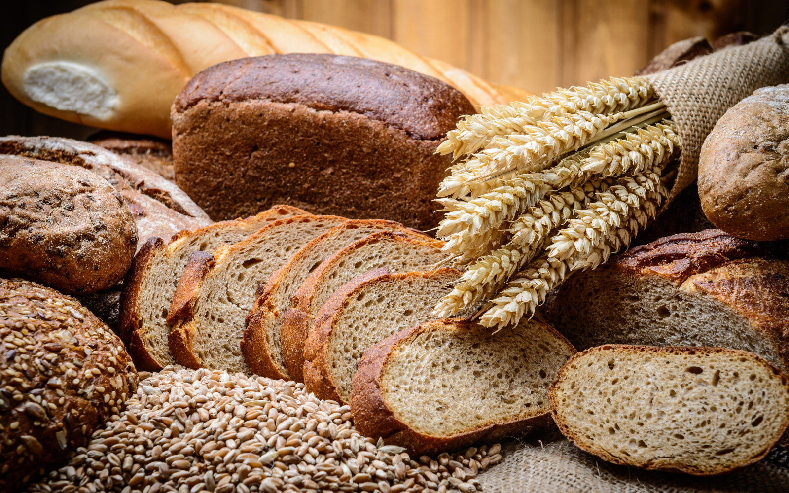 Various types of bread, wheat stalks, and grain on a rustic wooden surface.