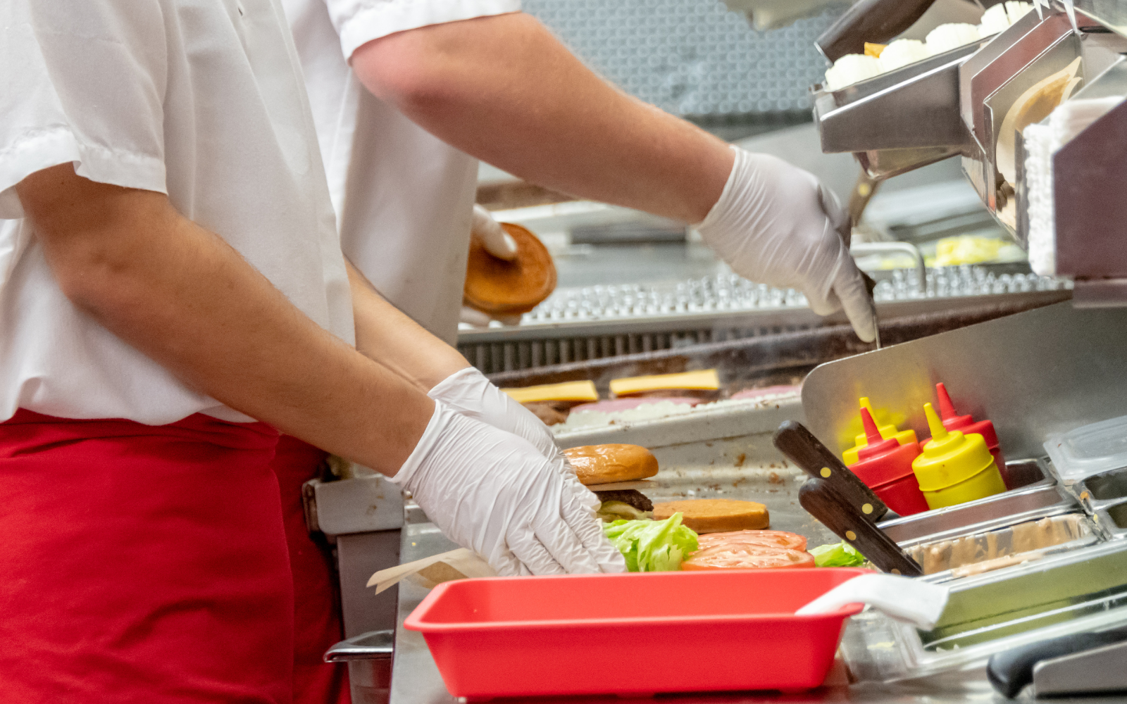 Two food workers wearing white shirts and gloves preparing sandwiches with lettuce and deli meat in a fast-food kitchen. Various condiments and ingredients are on the counter, including ketchup and mustard bottles, buns, and cheese.