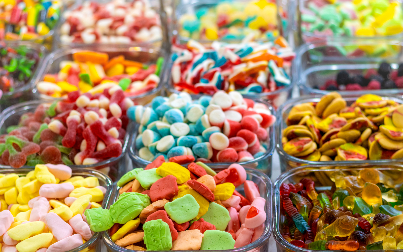 Multiple glass containers filled with colorful candies, including gummy worms, candy-coated chocolates, jelly beans, and various shaped sweets.