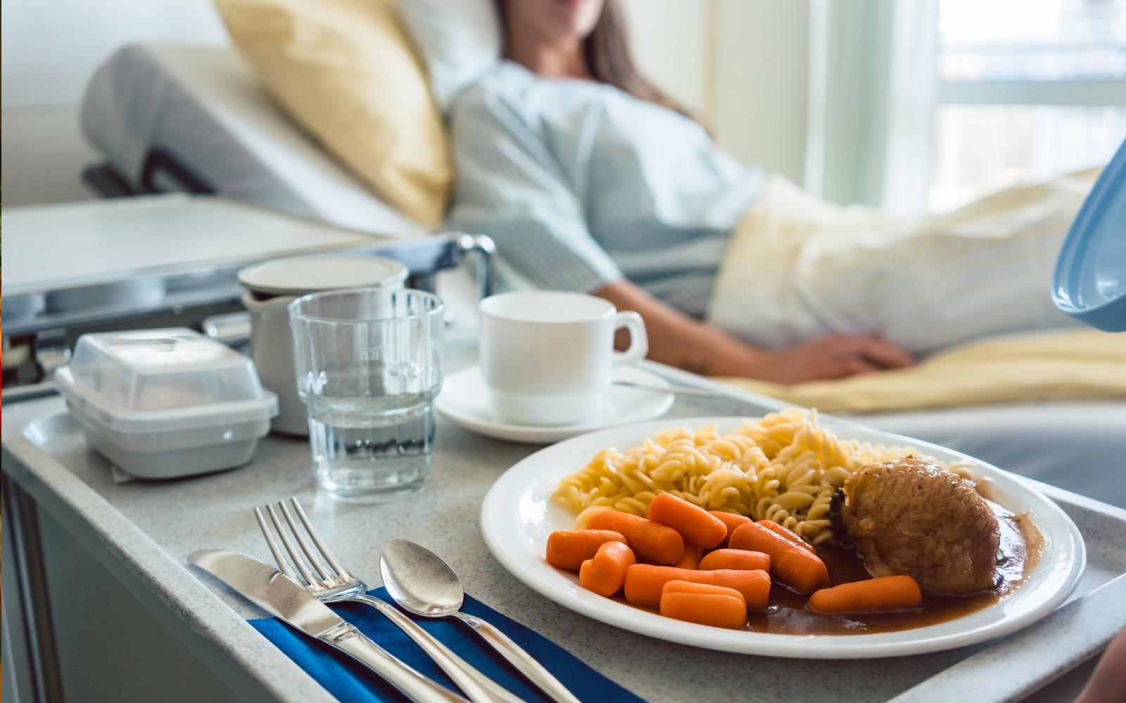 Hospital bed with an elderly patient, tray with food including carrots, noodles, and meatloaf, and a glass of water.