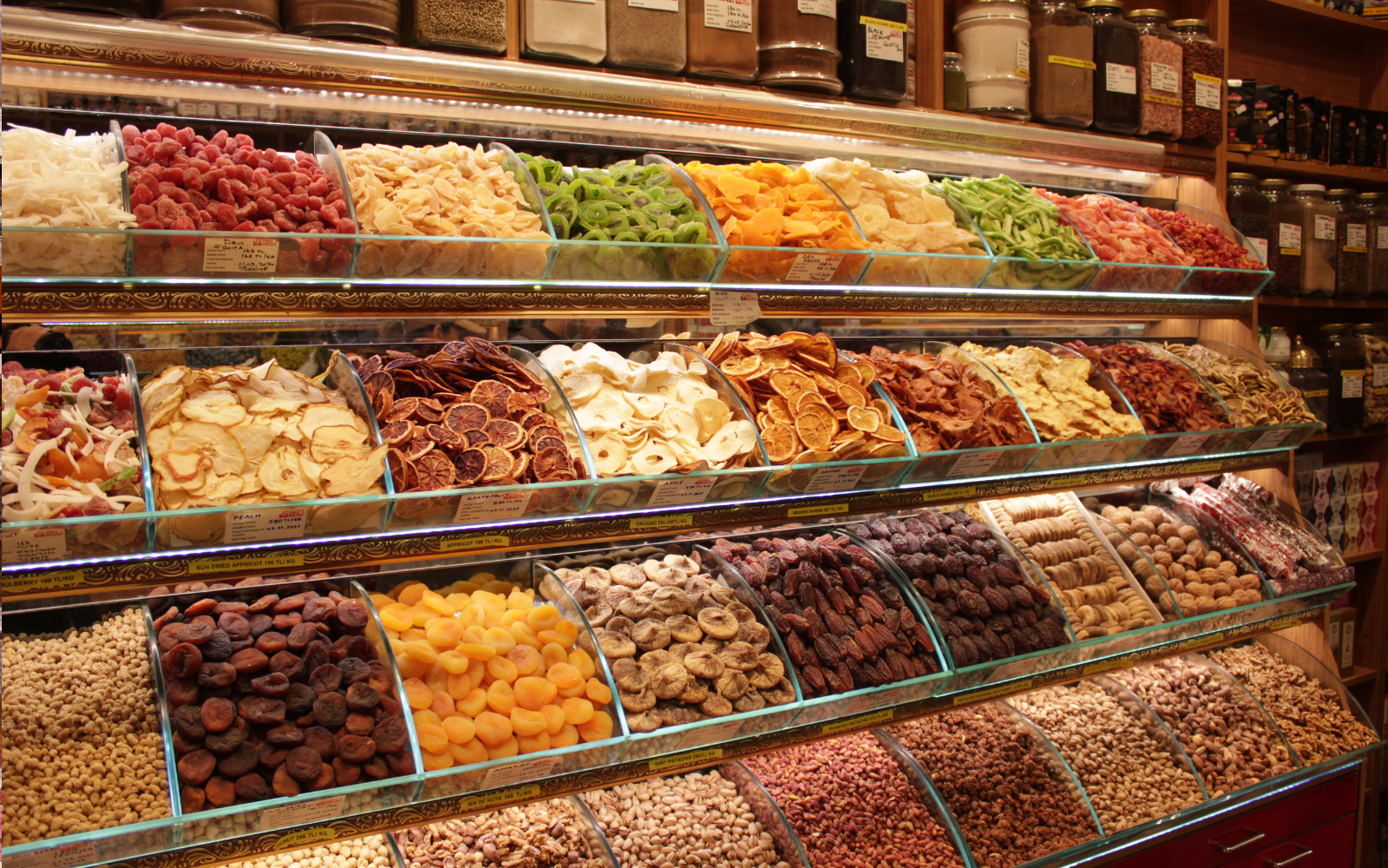 A display case filled with a variety of dried fruits and nuts arranged in rows, with spices in jars in the background.
