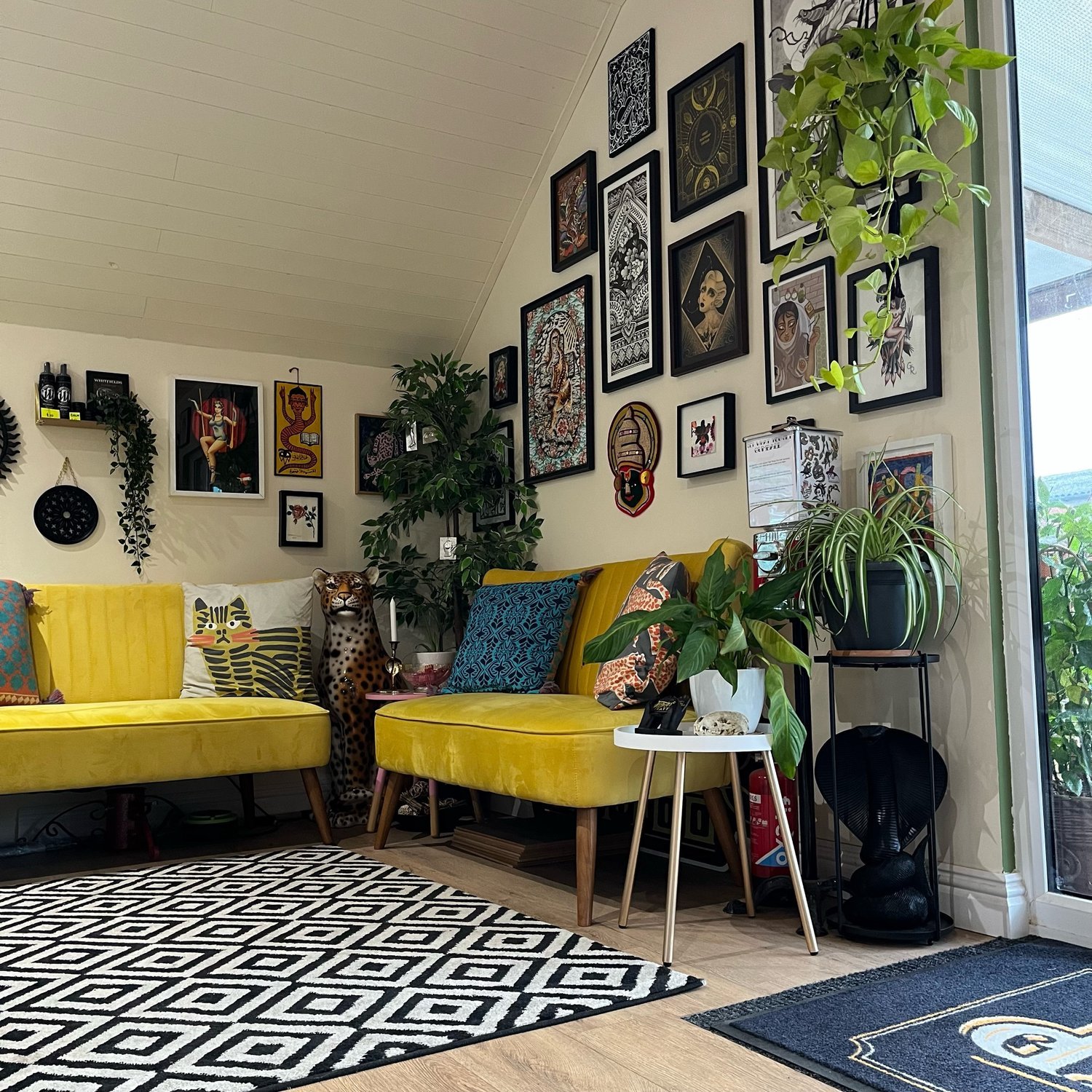 Eclectic living room with yellow couches, colorful throw pillows, a variety of framed artwork on the walls, and several green plants on tables and stands. A patterned black and white rug covers the wooden floor, and there's a decorative ceramic leopa