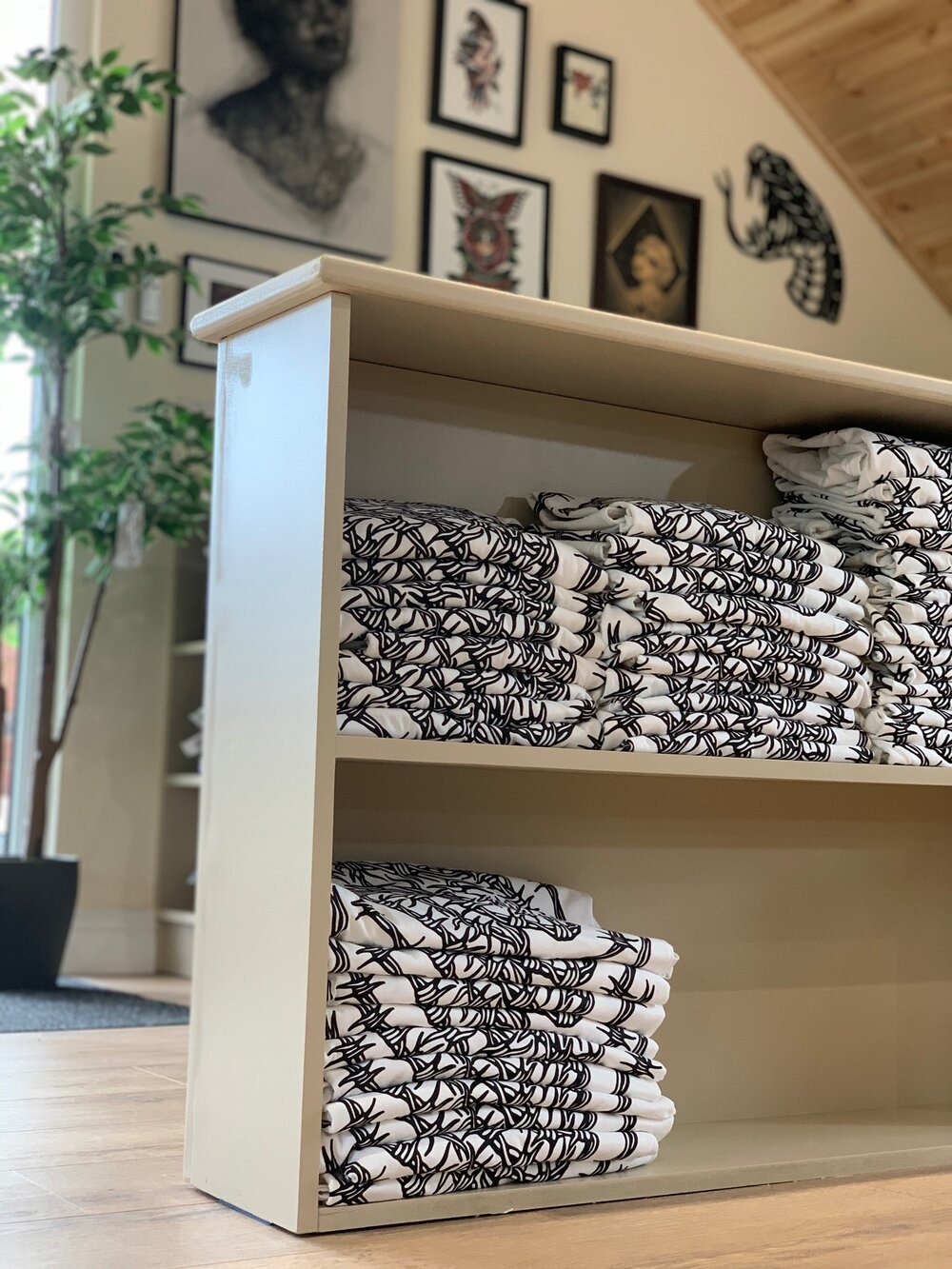 Shelves with folded white shirts featuring black patterns, displayed in an art gallery with framed artwork on the walls and a potted plant nearby.