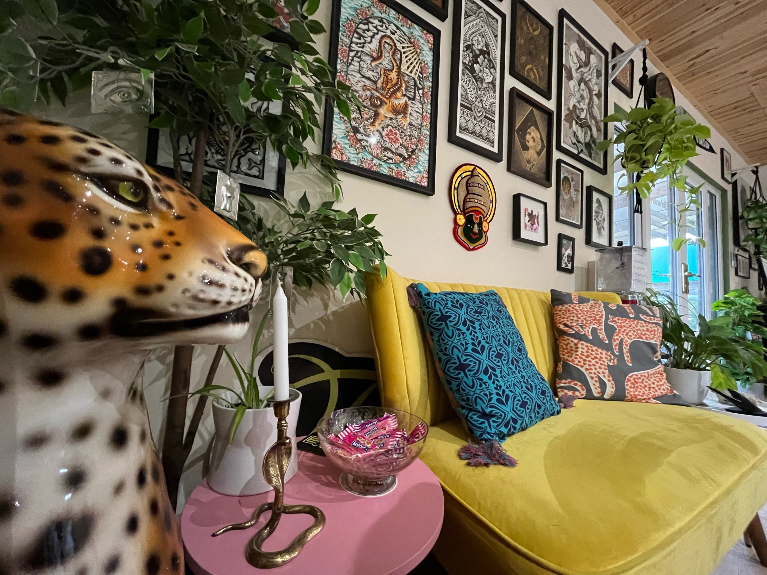 Colorful living room with leopard figurine, yellow sofa, blue and orange patterned pillows, gallery wall with various artwork, green plants, and snake-shaped candle holder.