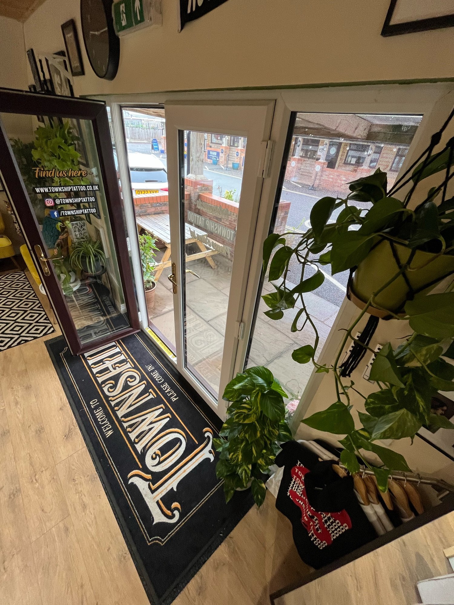 Interior of a tattoo shop with an open glass door leading outside. The floor has wooden flooring and a large black and white branded mat. Indoor plants are placed around the entrance, along with a clothing rack on the side. Outside, a street view is