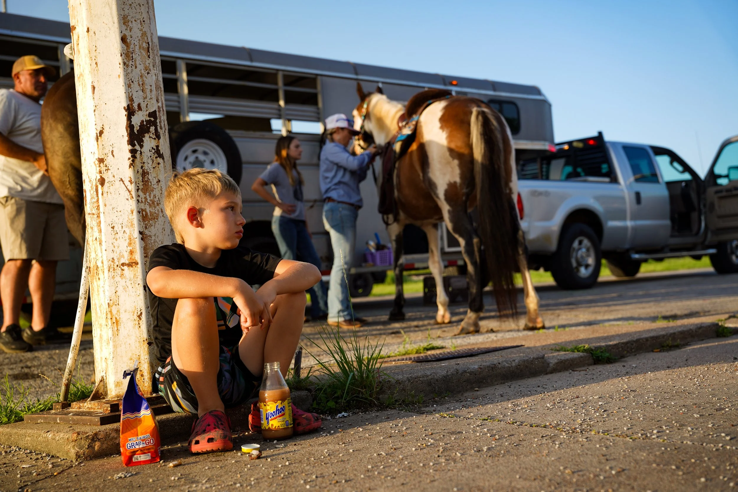 A young boy squats against a beam outside of a gas station in Millsap, Texas while family members ready their horses for the annual Parker County Sherriff's Posse "Bringin' In the Herd" Cattle Drive June 6, 2025.