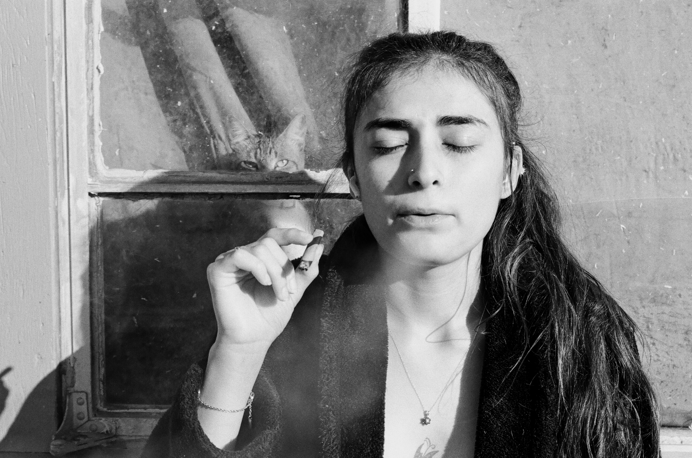 A young woman with long hair, eyes closed, smoking weed outside a window. A cat is peeking through the window behind her.