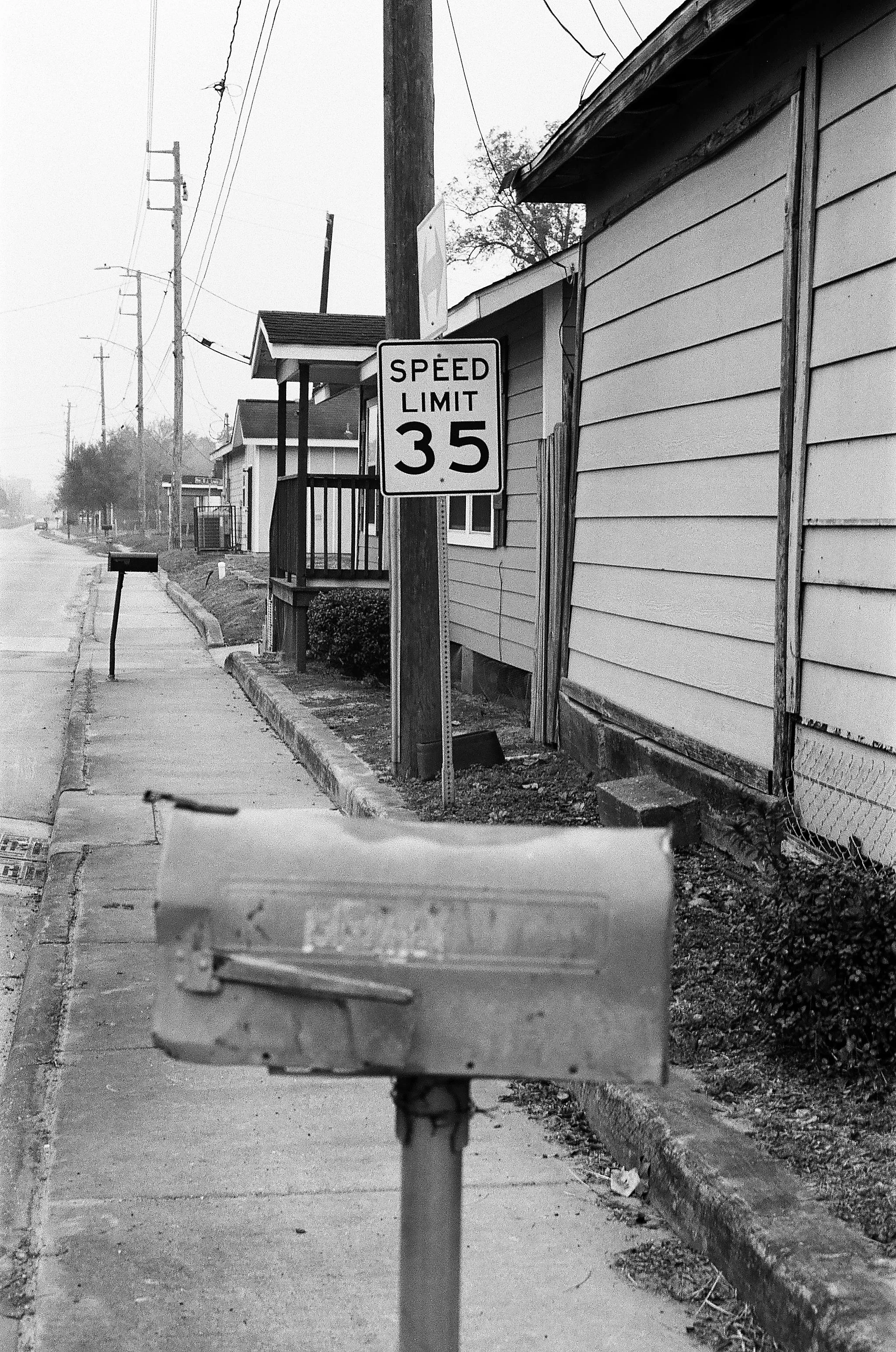 A black-and-white photo of a sidewalk with a speed limit sign showing 35 mph, a mailbox, and residential houses with overhead power lines.