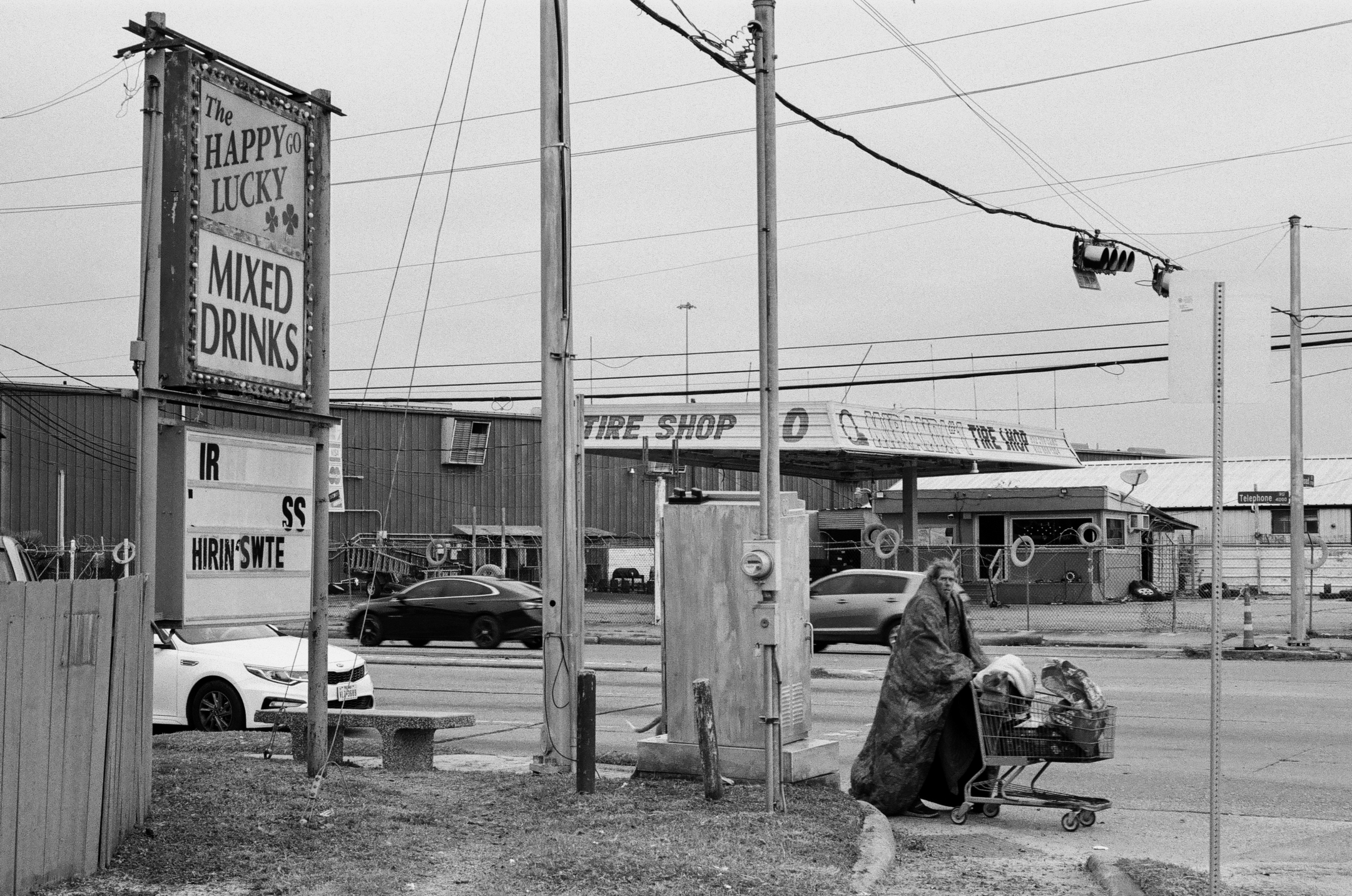 A man in a coat stands by a shopping cart on a sidewalk next to a street, with cars driving by. There are utility poles with wires, a sign for a tire shop, and another sign advertising mixed drinks. The scene appears to be in an urban or semi-industrial area, captured in black and white.