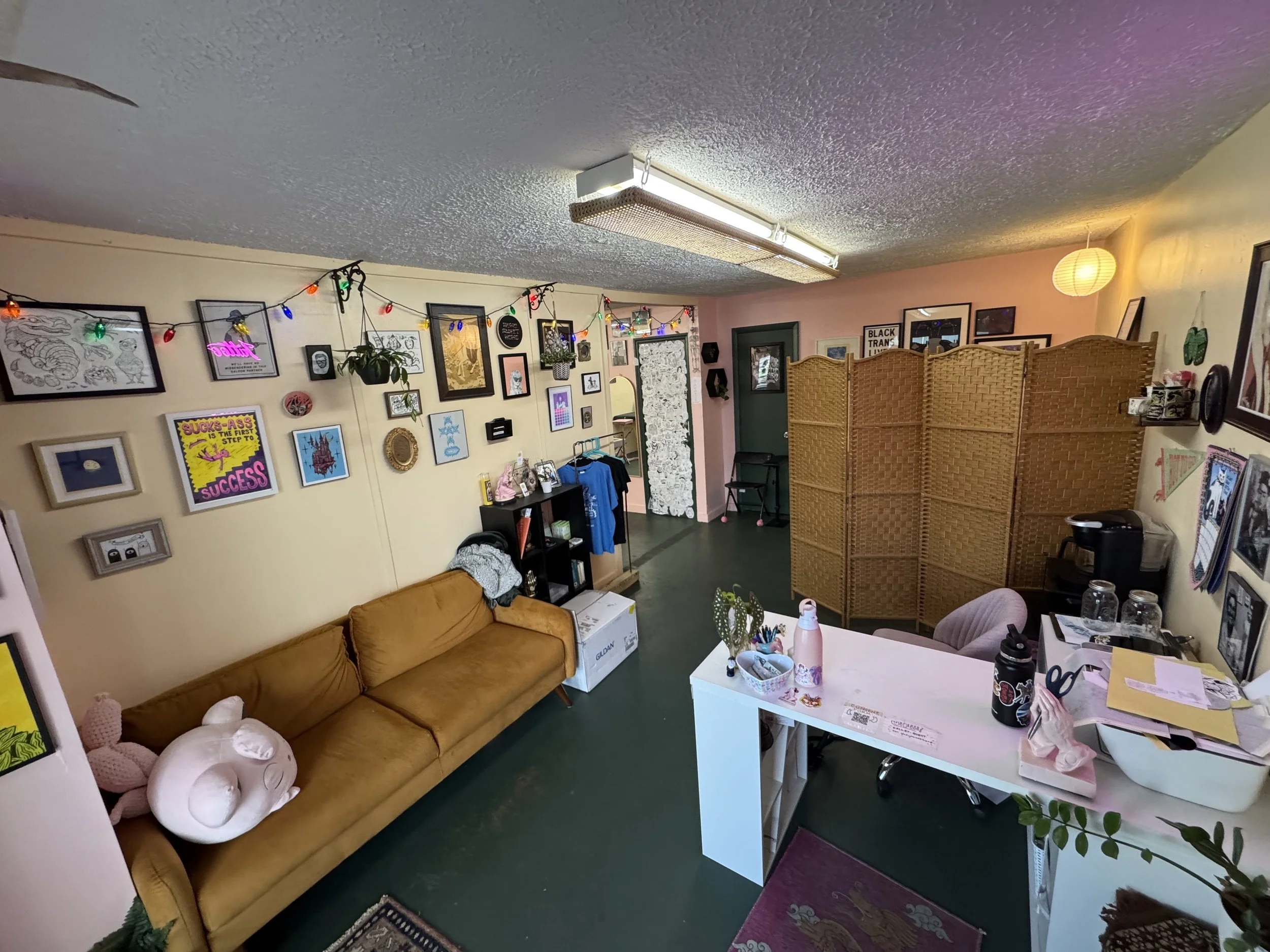 The image shows a cozy room with various wall decorations, including framed pictures and string lights. There's a mustard-colored couch with plush stuffed animal toys on it. A white table with pink bottles, papers, and miscellaneous items is in the foreground. A woven room divider stands against the back wall, and there's a plant and other furniture throughout the room.