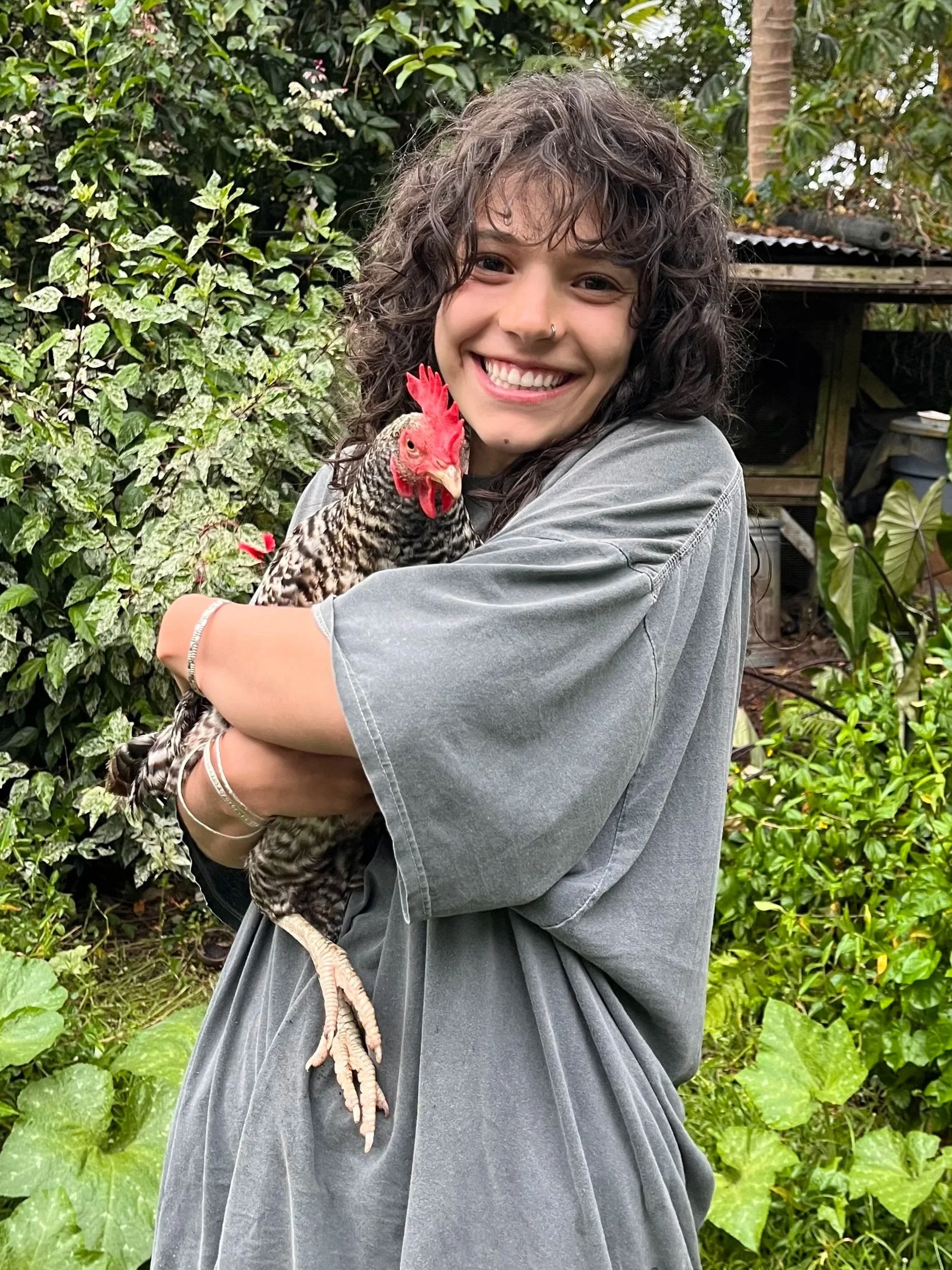 A smiling woman with curly hair holding a chicken in a lush garden.