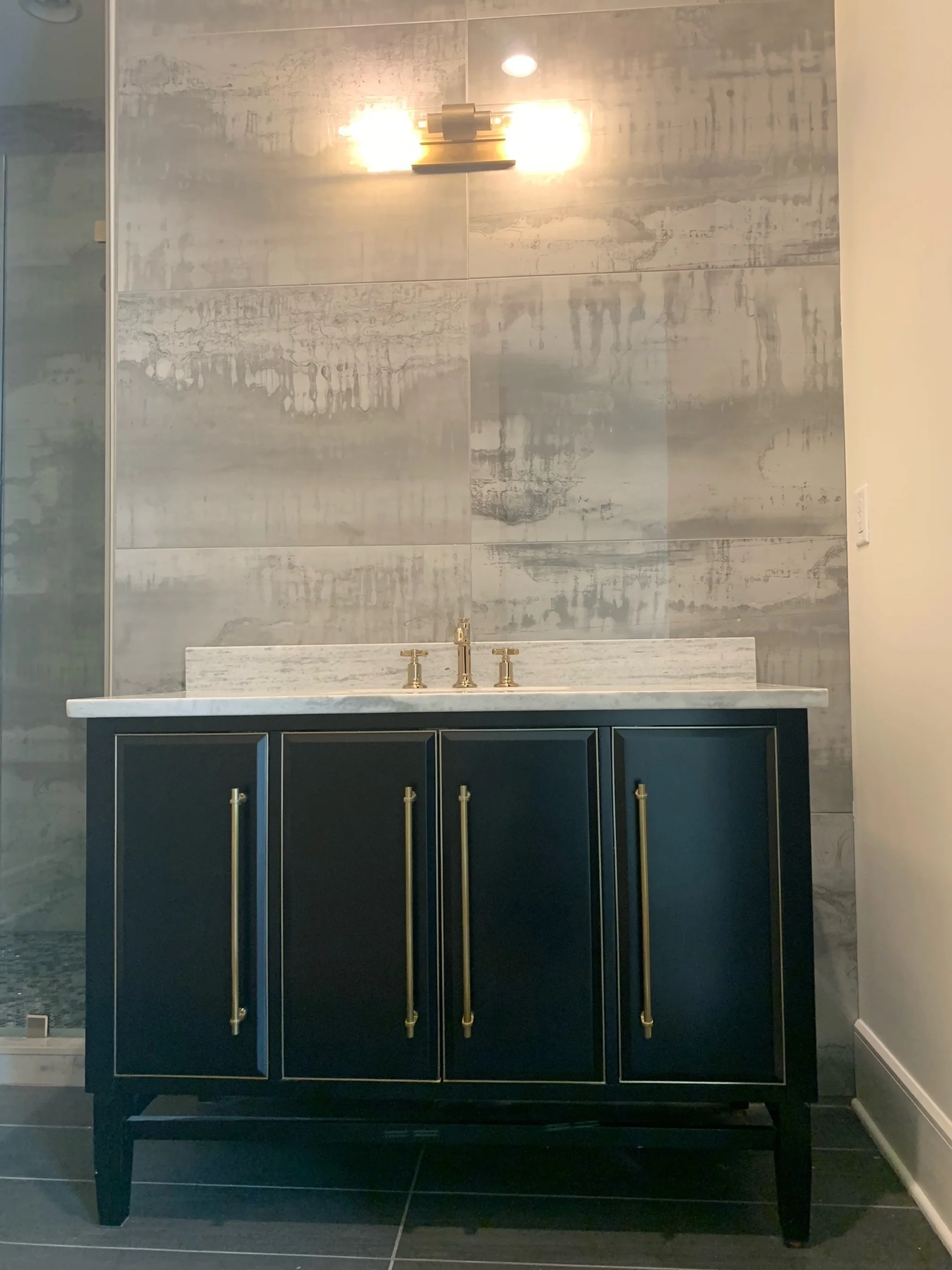 Bathroom vanity with dark blue cabinets, gold handles, white marble countertop, gold faucet, and gray tiled wall behind.