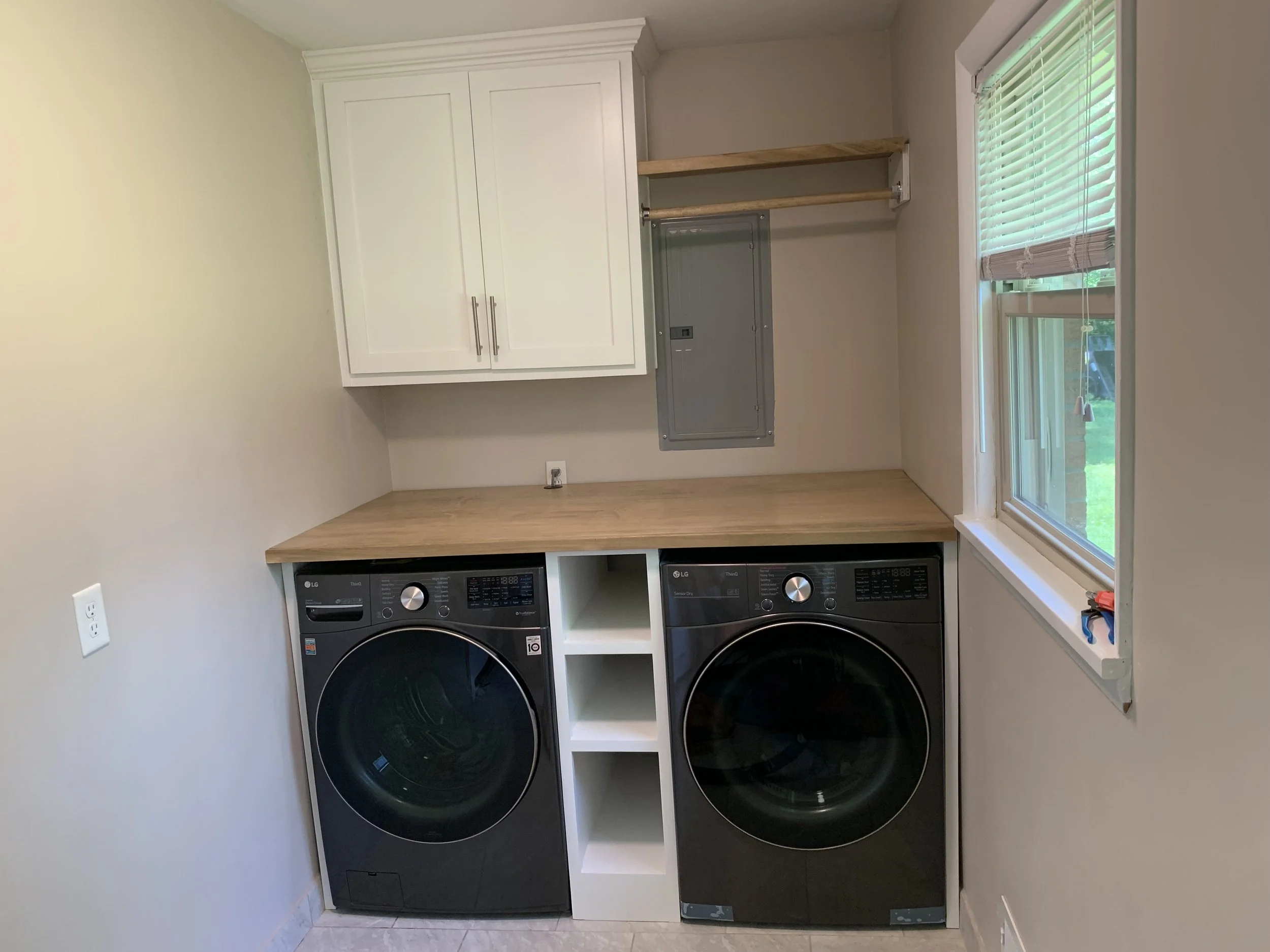 Laundry room with a black front-load washer and dryer, white upper cabinets, a wooden countertop, and a window with blinds.