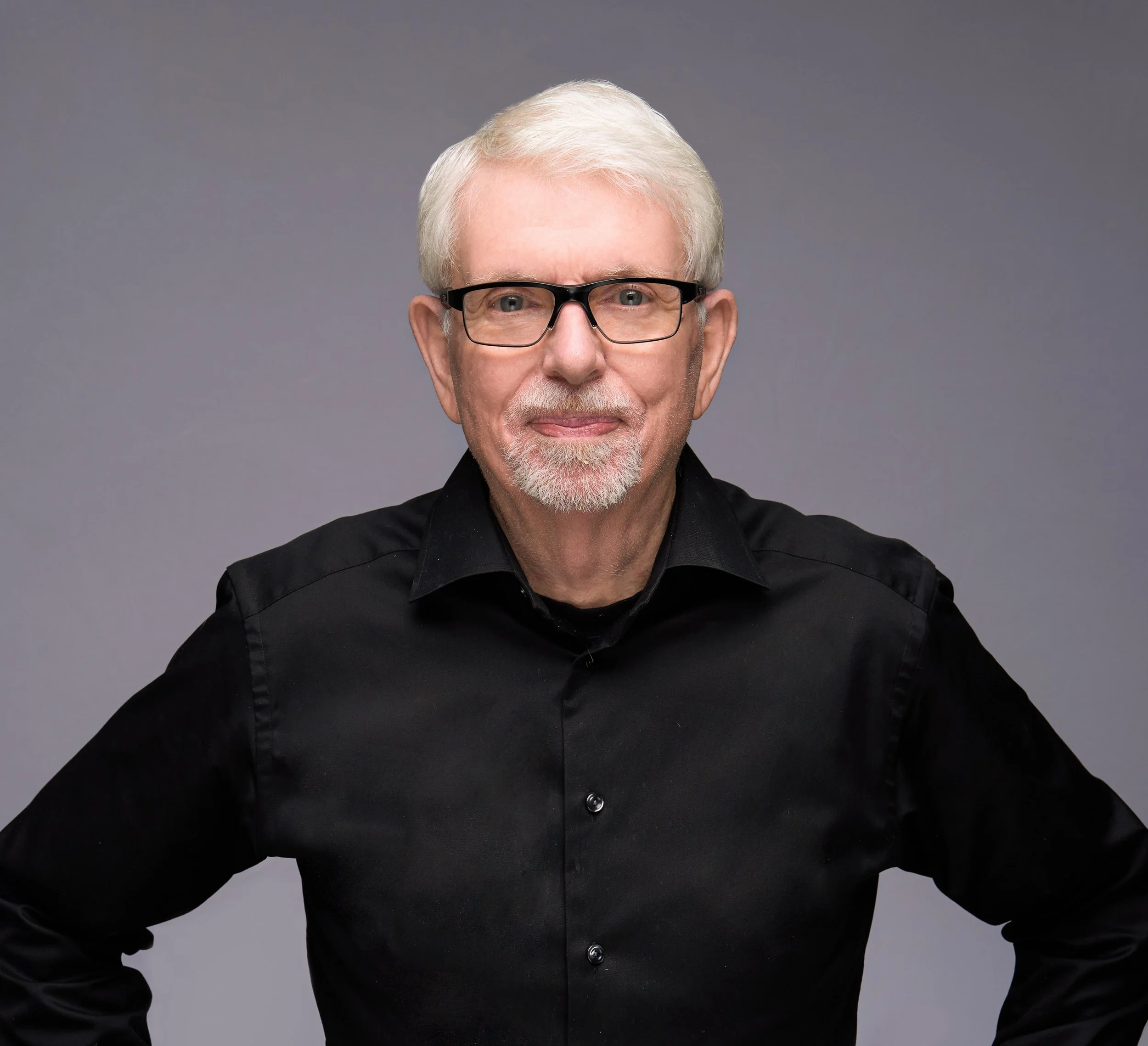A portrait of an older man with white hair, glasses, and a beard, wearing a black button-up shirt, standing against a gray background.