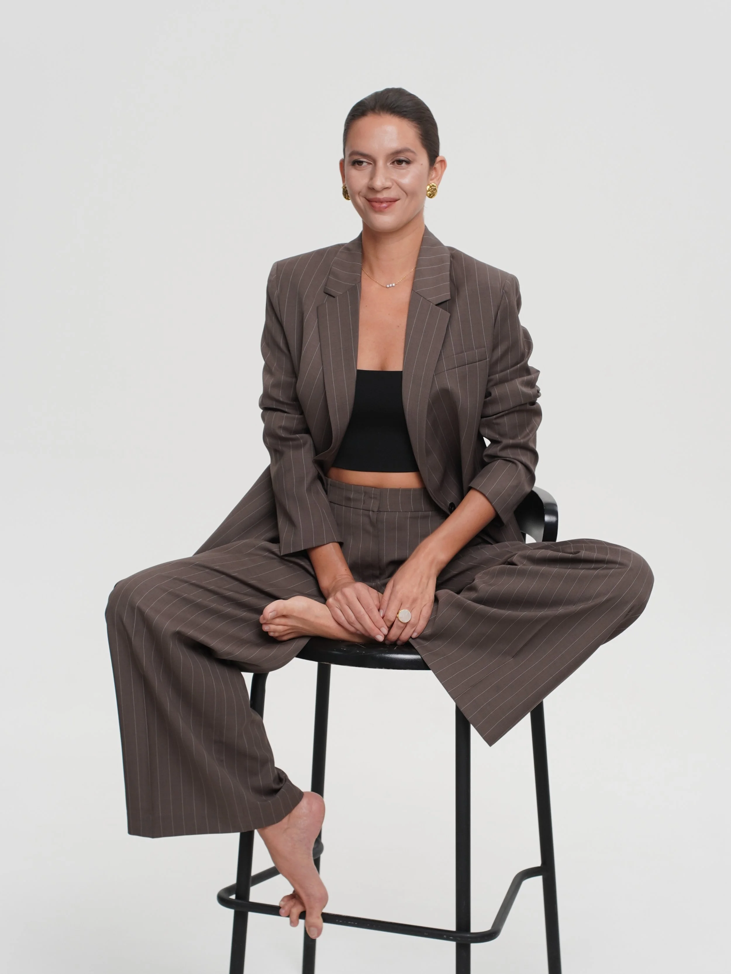 A woman in a brown pinstripe suit with a black crop top, sitting cross-legged on a black stool against a plain white background, smiling gently.