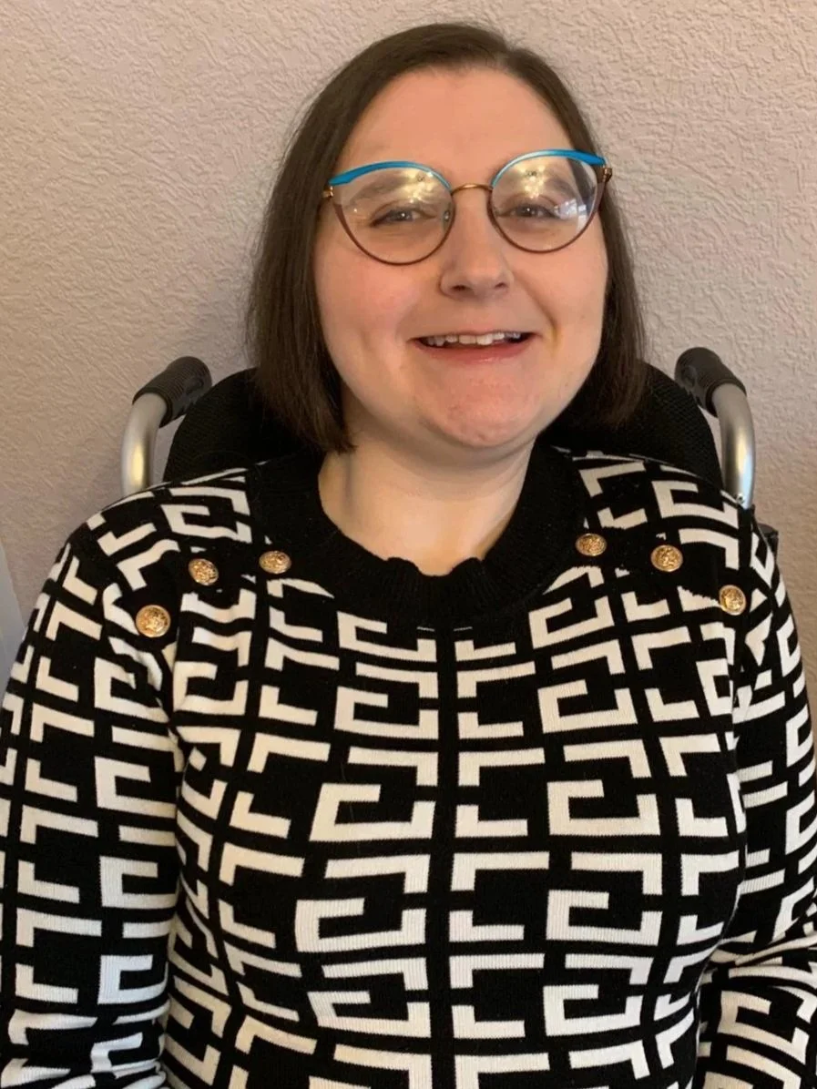 A woman with short brown hair and glasses sits in a wheelchair, wearing a black and white patterned sweater, smiling at the camera.