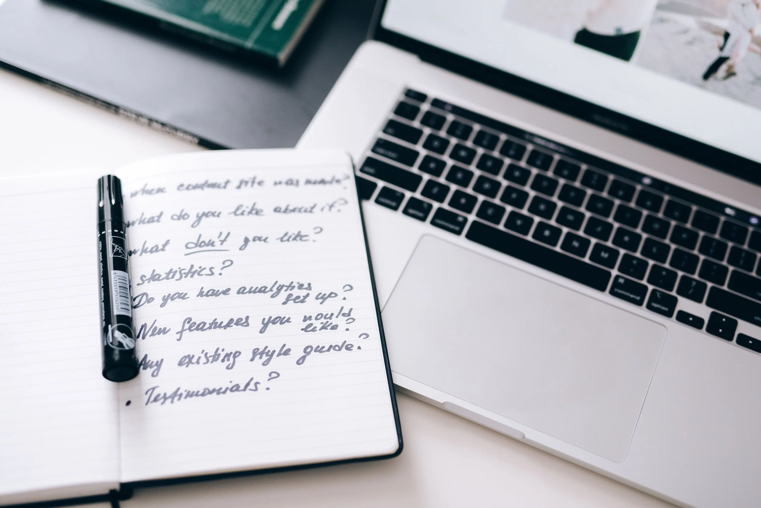 Open notebook with handwritten notes, a black pen on top, and a laptop nearby on a white surface.
