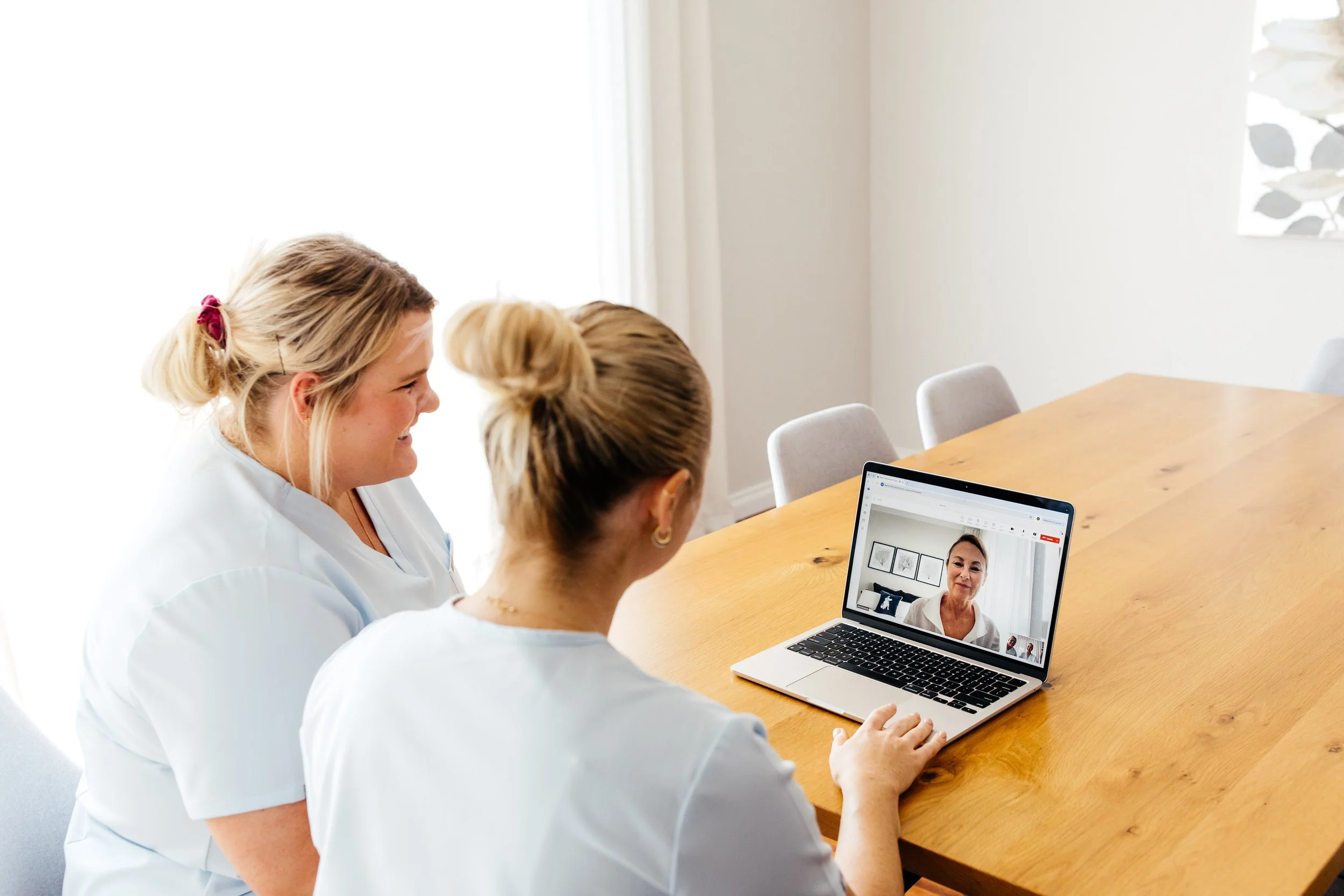 Two women in scrubs sitting at a table, video conferencing with another person on a laptop.