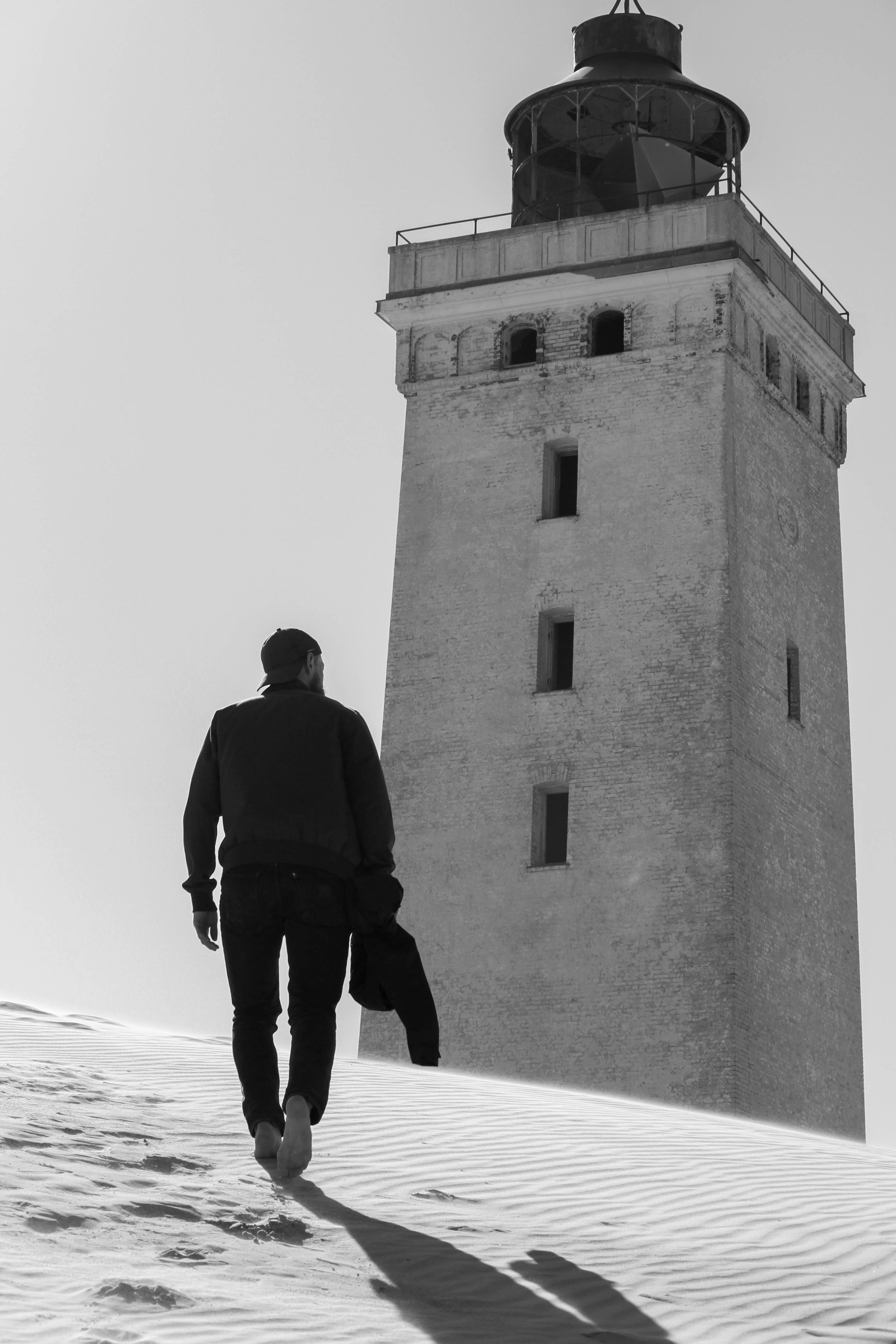 A lone figure walking toward a pale lighthouse across snow, black and white.