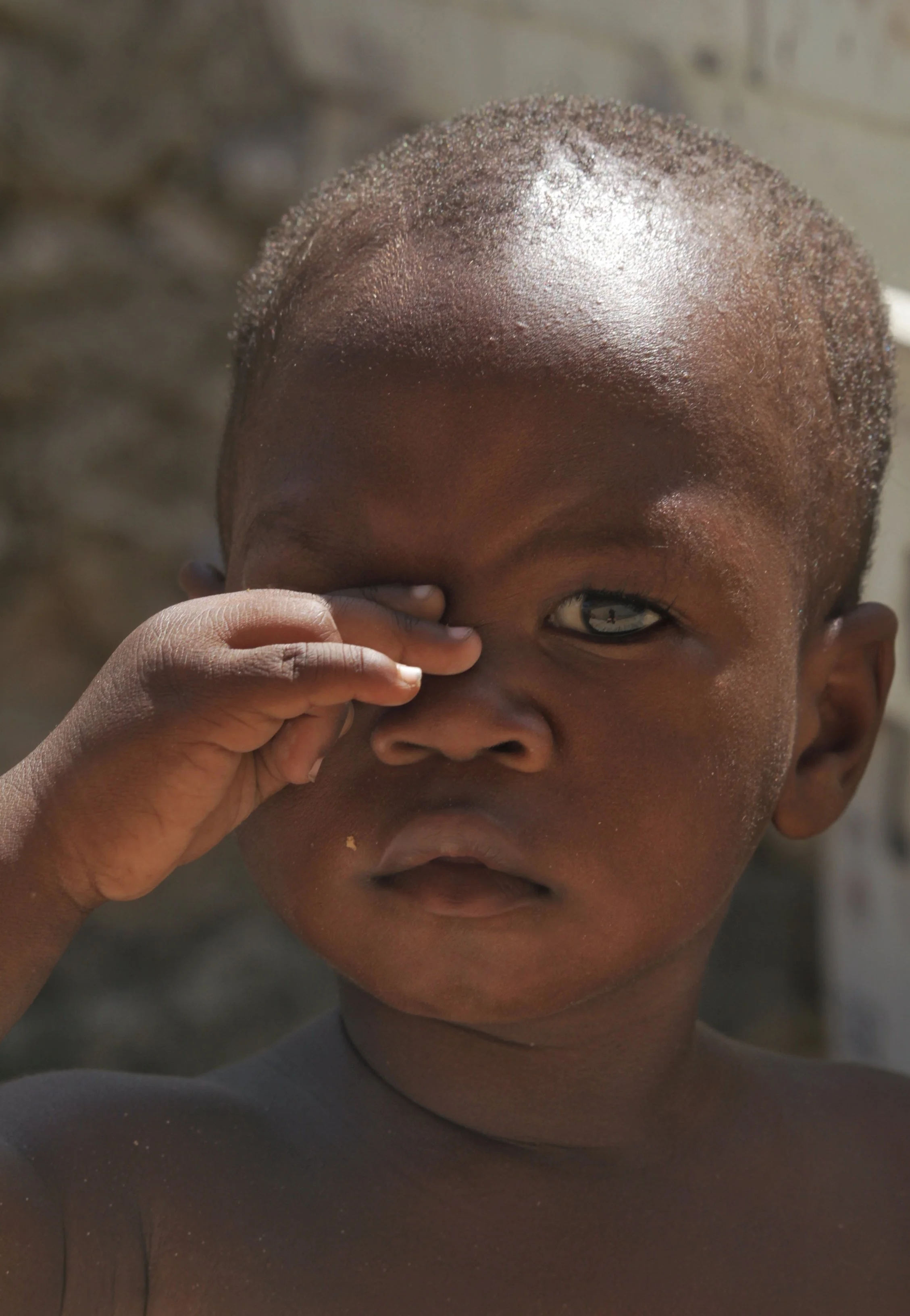 A young Zanzibari child raising a hand to one eye, looking directly at the camera.

