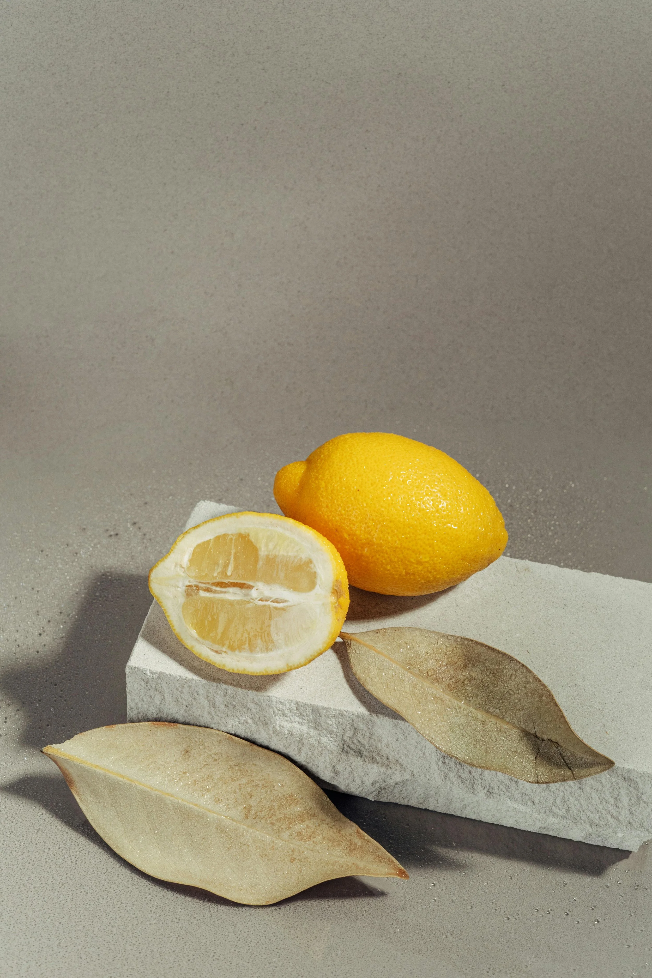 Lemon on a white textured platform, with dried leaves, against a gray background.