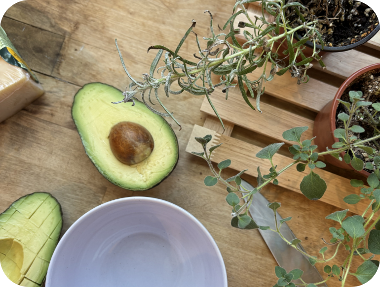 Half an avocado with seed, sliced avocado, potted herbs, and a white bowl on a wooden surface.