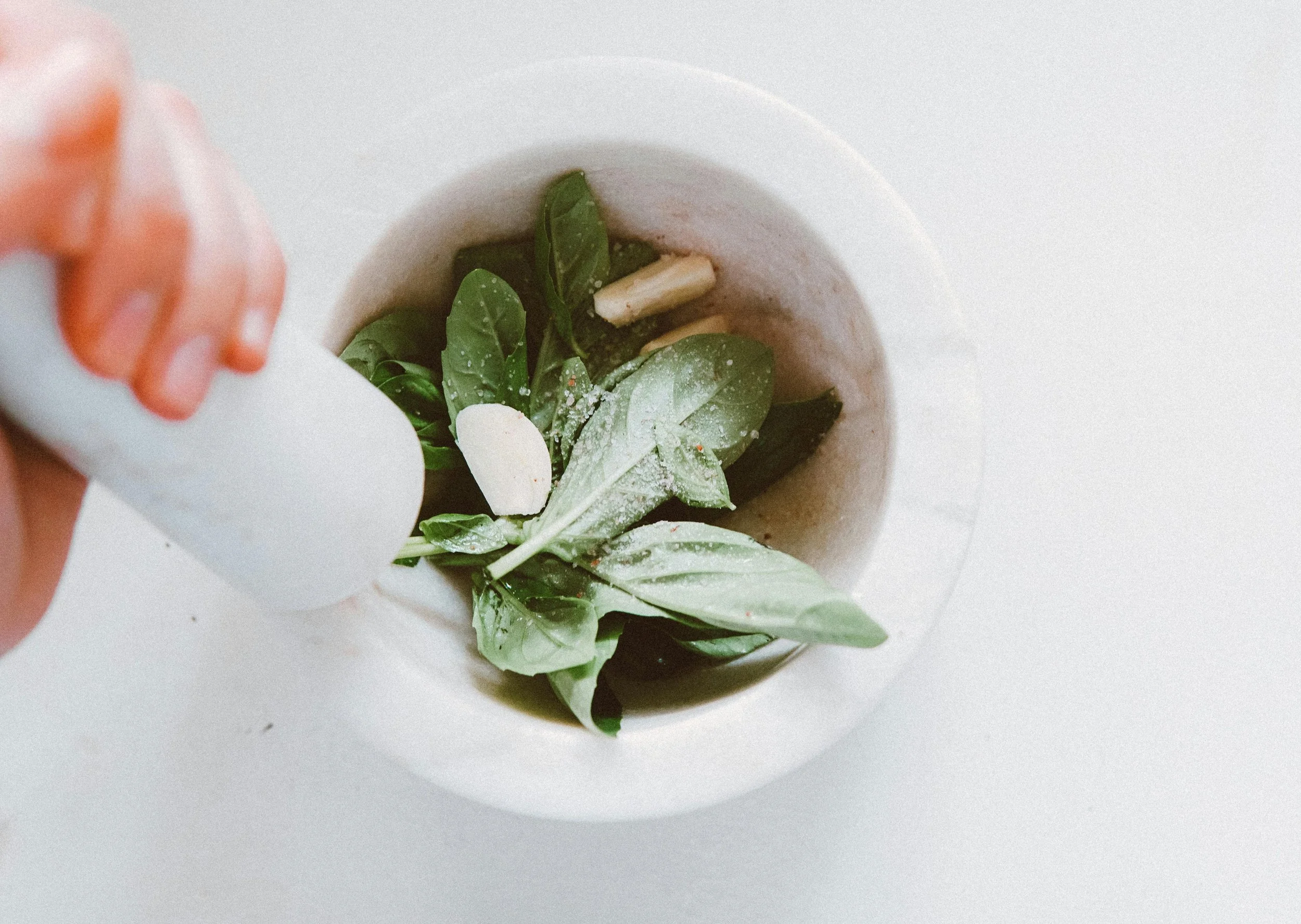 Person adding salt to a mortar filled with fresh basil leaves and garlic cloves.