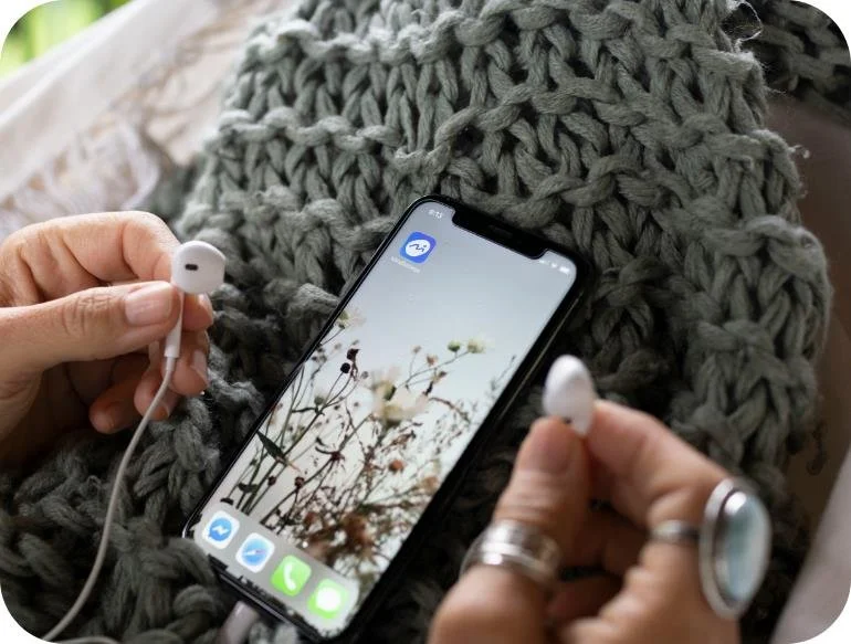 Person holding white earbuds and an iPhone with a nature scene on the screen, sitting on a chunky gray knit blanket.