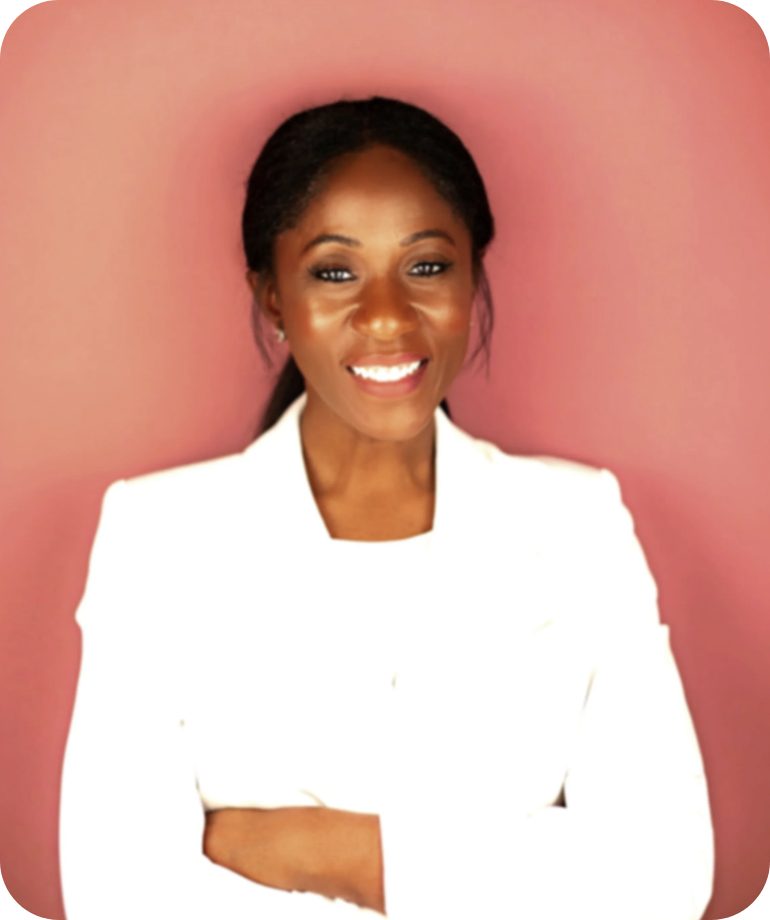 A woman with dark hair in a ponytail, wearing a white blazer, smiling with arms crossed against a pink background.