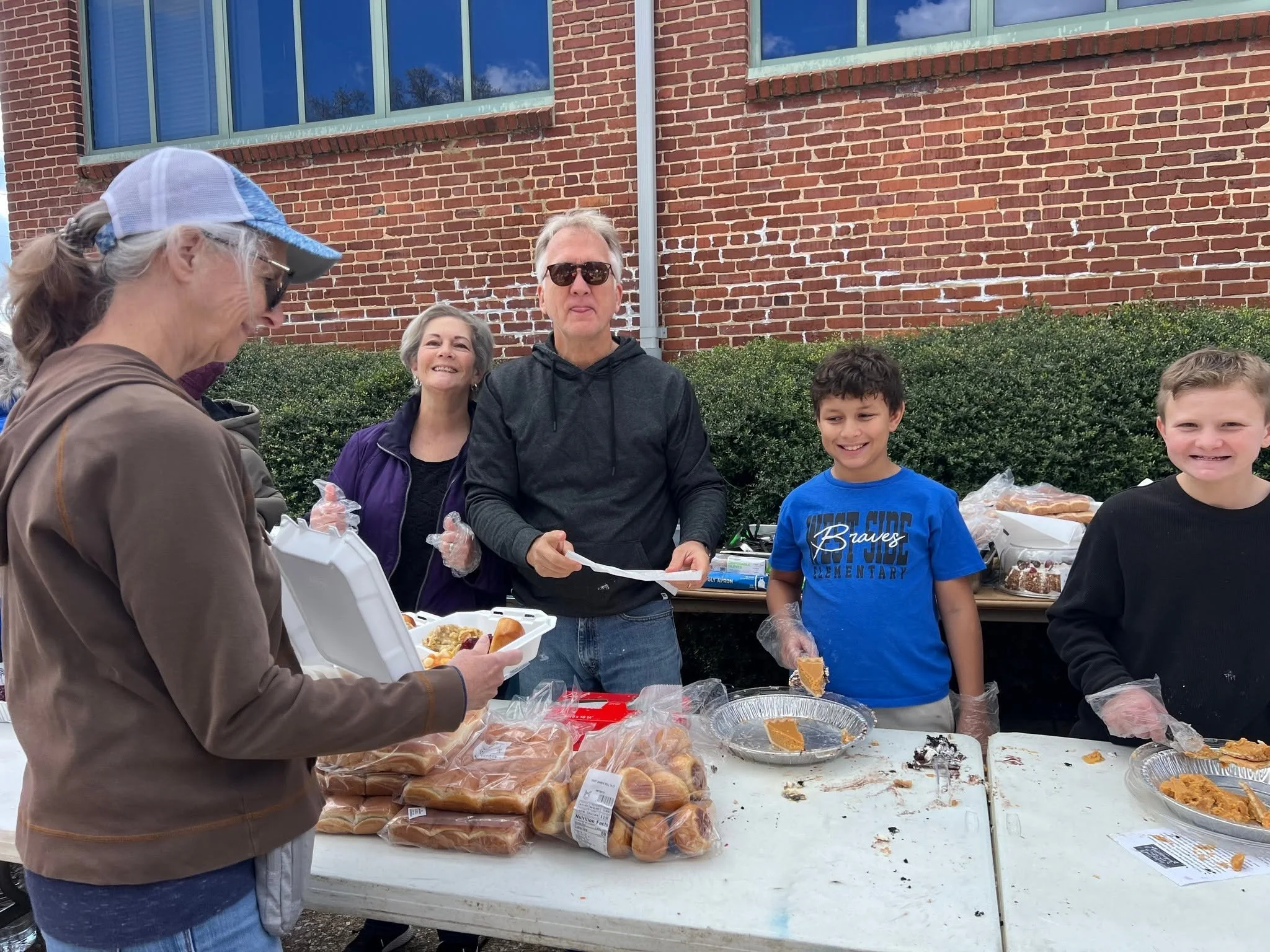 People standing in front of a brick building with large windows, scattering food and supplies during an outdoor community event.