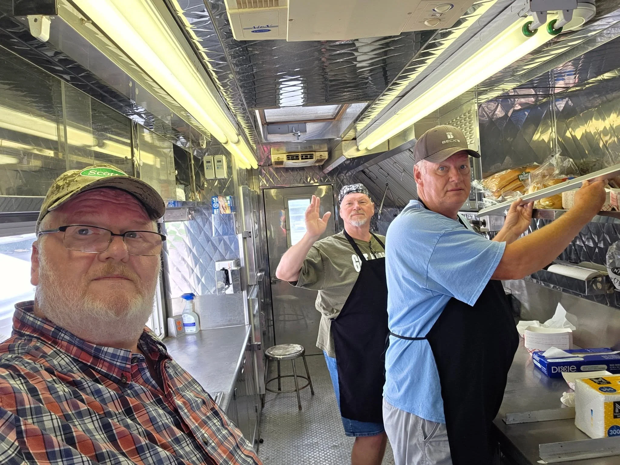 Inside a food truck with three men, two of whom are facing viewer and one facing the food prep area, all appear to be staffing the truck. The man in the foreground is wearing glasses and a plaid shirt, the man in the middle is wearing a bandana and an olive green apron, and the man on the right is wearing a baseball cap and a blue shirt. The truck has reflective metal surfaces, fluorescent lighting, and shelves with food items and supplies.
