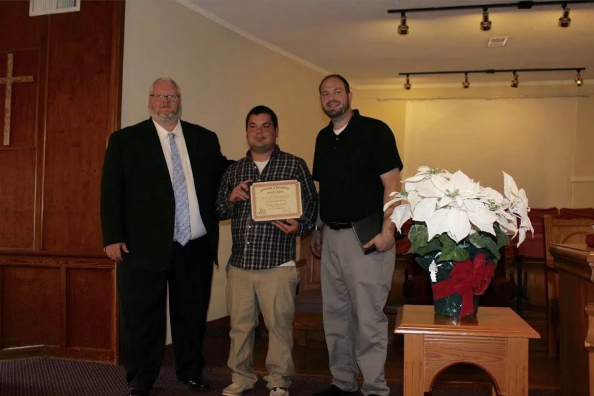 Three men standing together in a room, one man is holding a certificate, a poinsettia plant is on a table in front of them, and a cross and church-like wood paneling are visible on the wall behind them.