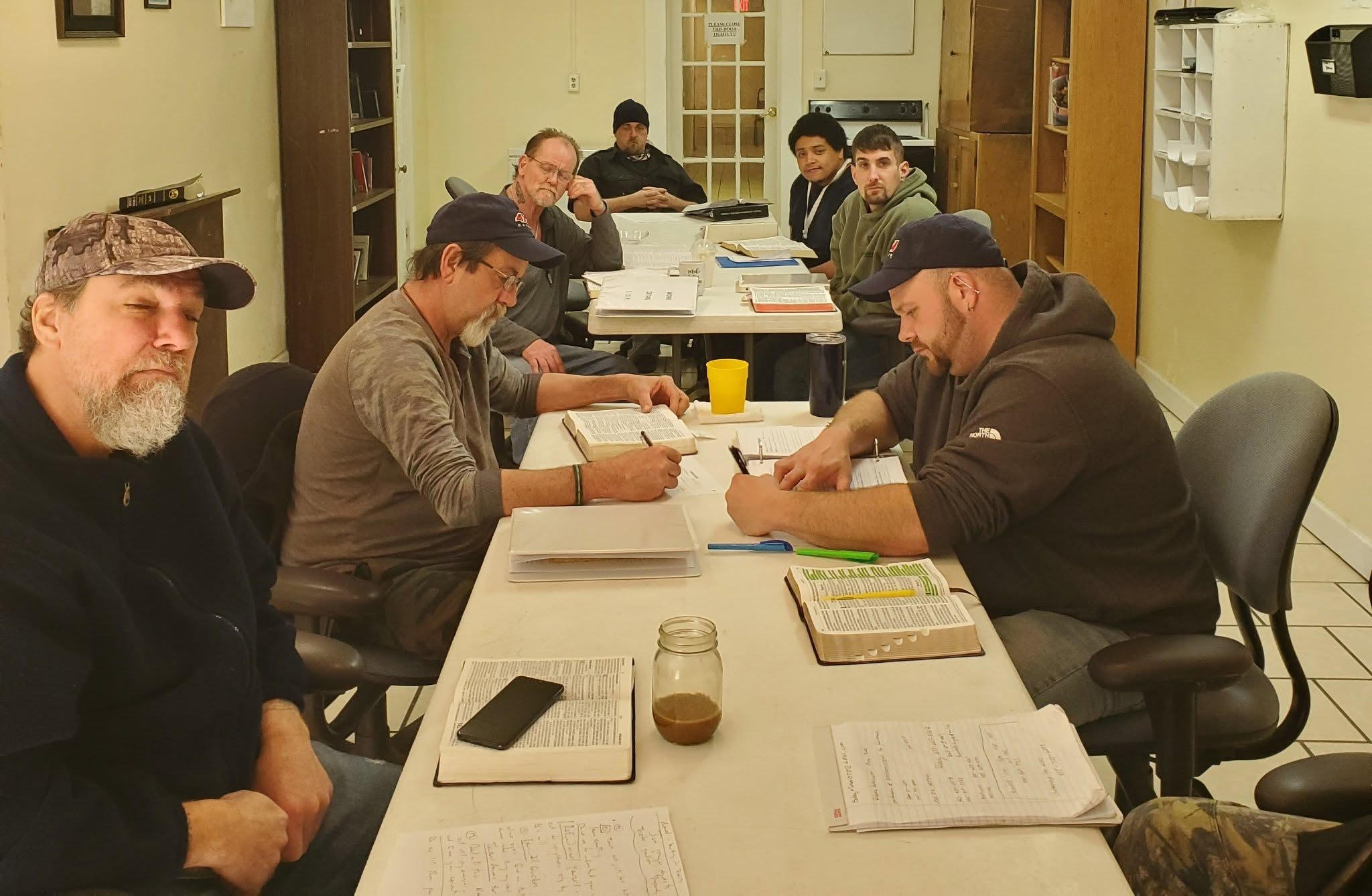 A group of seven men sitting at a rectangular table in a room, appearing engaged in studying or discussion with open books, notebooks, and writing materials on the table.
