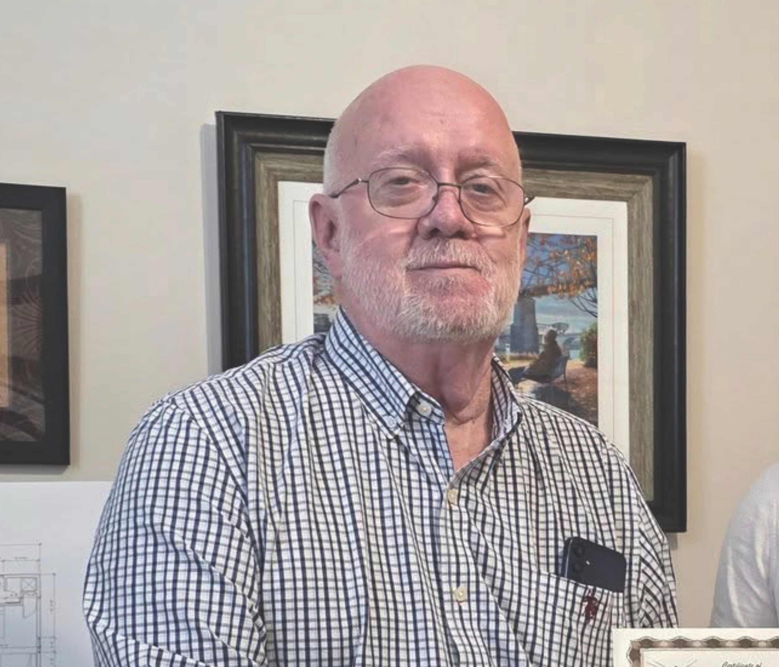 A smiling elderly man with glasses, a bald head, a white beard, and mustache, wearing a checkered shirt, standing in front of framed artwork on a beige wall.