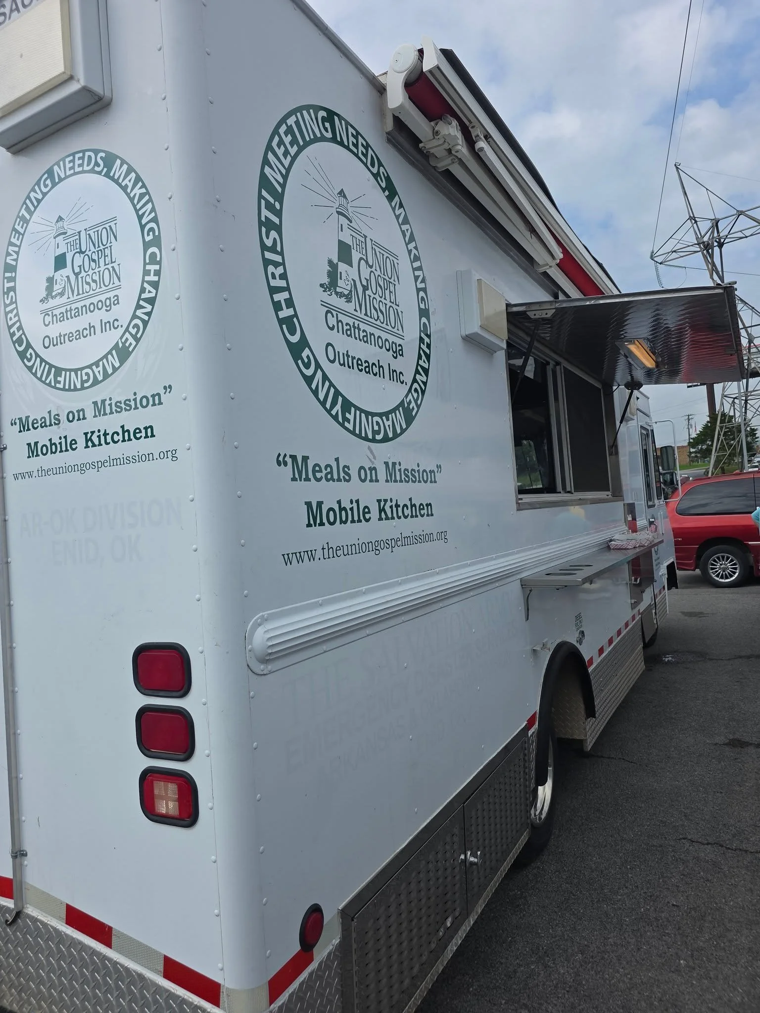 A white mobile kitchen truck with logos of The Union Gospel Mission Chattanooga Outreach Inc., a lighthouse graphic, and text indicating it is for meals on mission. The truck has a serving window and is parked next to other vehicles under a partly cloudy sky.