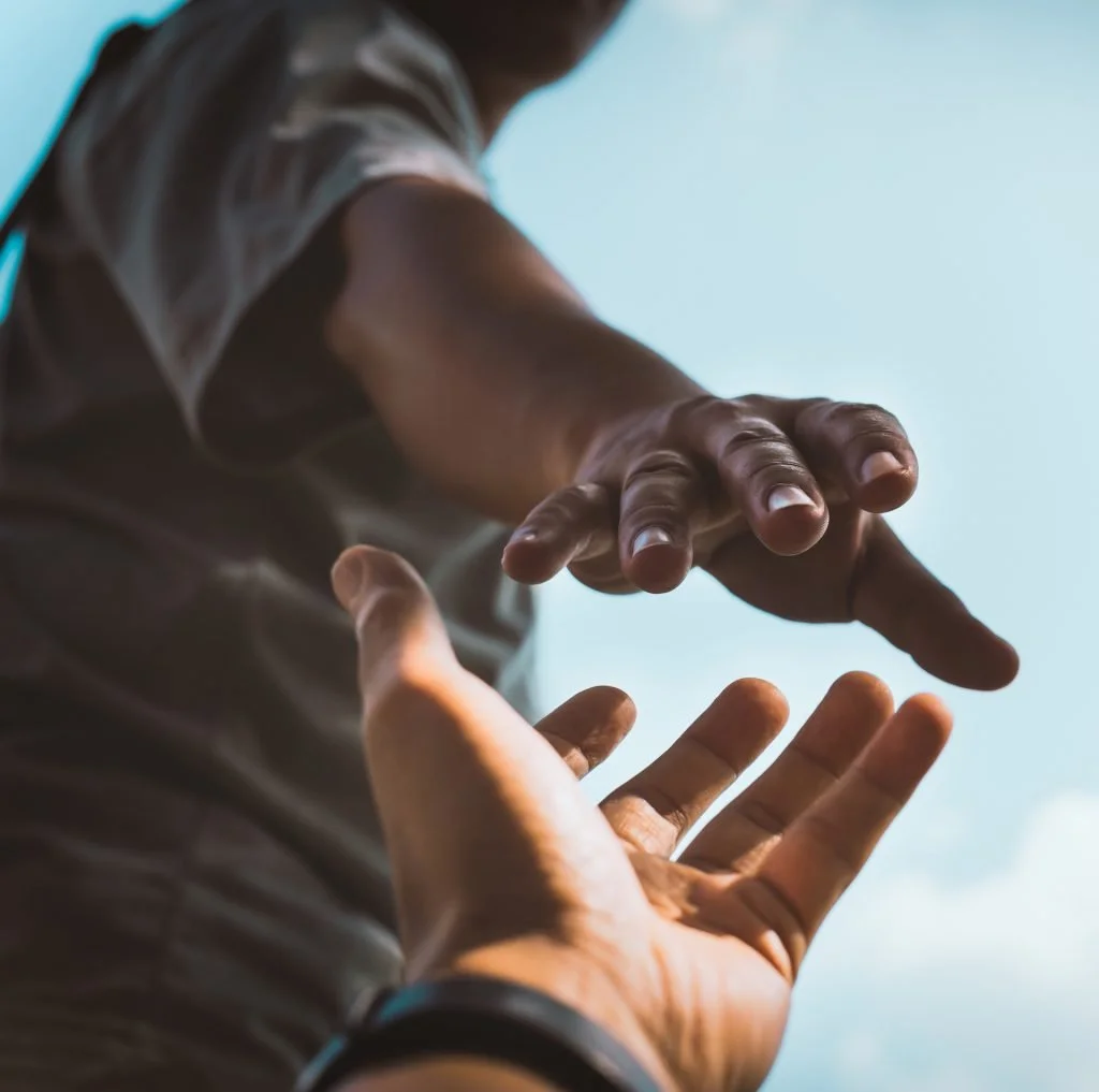Two people reaching out their hands towards each other, one with darker skin and one with lighter skin, against a bright sky background.