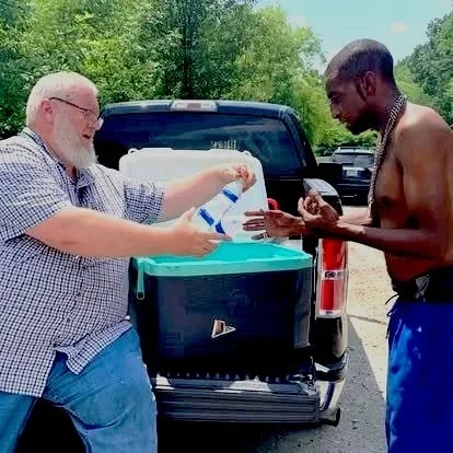 A man with white hair and glasses is handing a bottle to a shirtless man with dark skin and a necklace, standing beside a truck with the tailgate down, outdoors in a parking lot with trees in the background.
