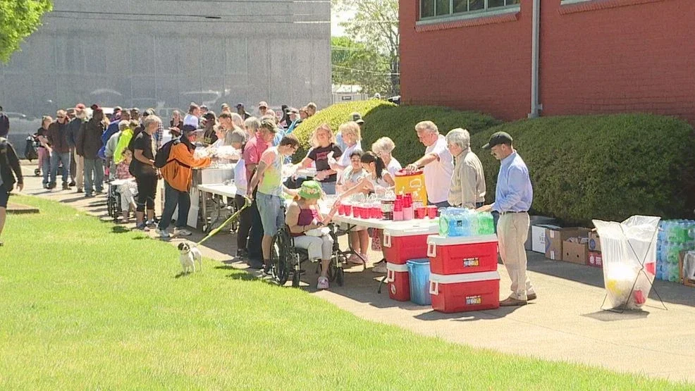 Long line of people waiting outside near a food and drink table on a sunny day, with supplies and coolers on the ground.