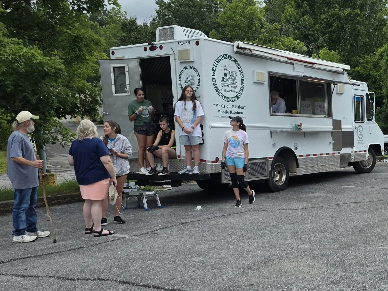 A mobile kitchen truck with the logo and information for Union Gospel Mission in Chattanooga. Several young people are gathered around and on the truck, with some engaged in conversation and others sitting or standing. There is a man with a cane talking to a woman near the truck.