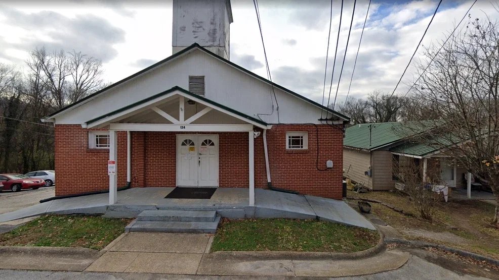 A small brick church with a white front porch and double doors, set on a slightly elevated sidewalk with steps. There are cars parked nearby and wires overhead, with trees and neighboring houses in the background.