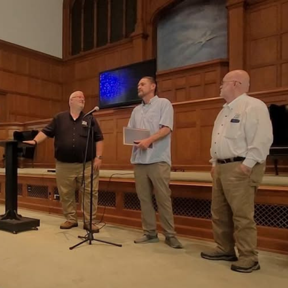 Three men standing in front of wooden paneling, with one speaking into a microphone, in a room with a raised platform and a large screen behind them.