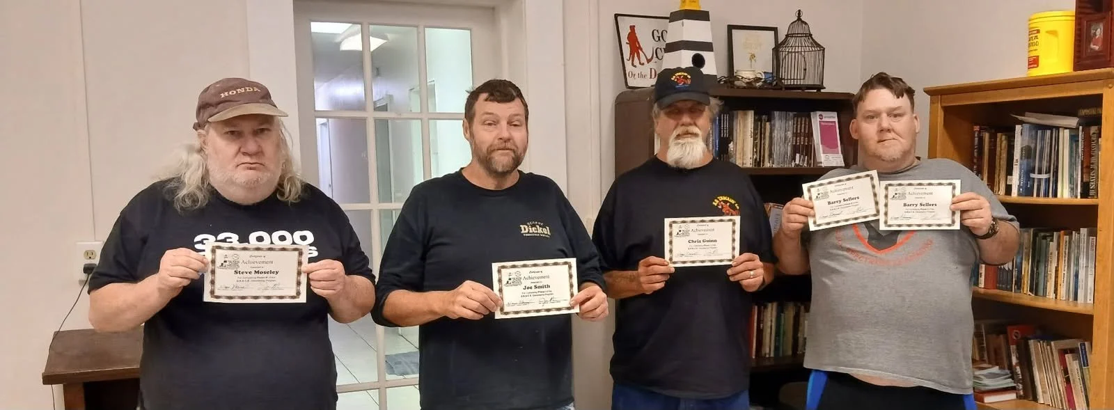 Four men standing in a room holding certificates. The room has bookshelves and framed pictures in the background.