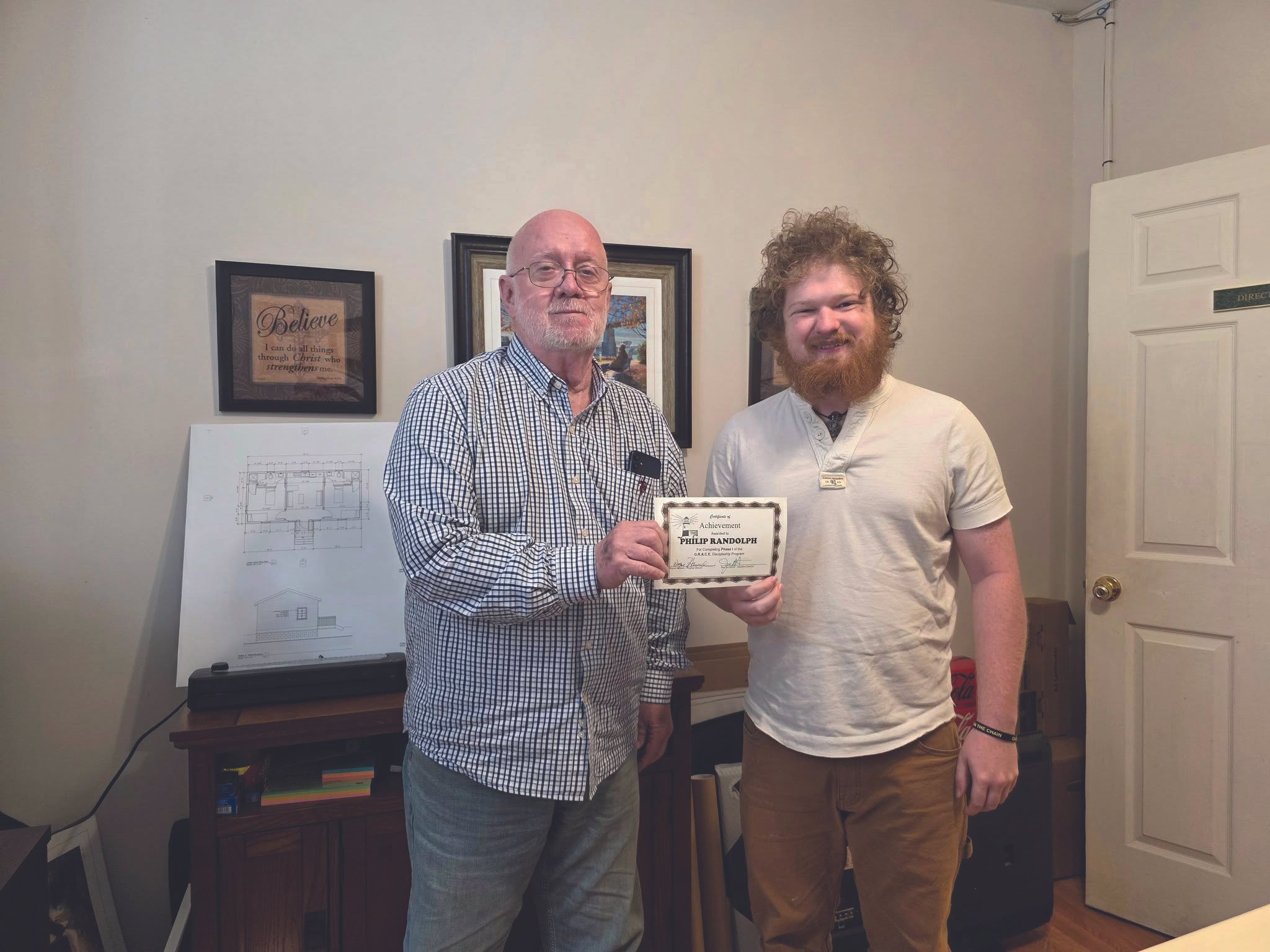 Two men standing inside a room, with one holding a certificate of achievement. The man on the left is bald with glasses, wearing a checked shirt, and the man on the right has curly hair and a beard, wearing a light-colored shirt. There are framed pictures and architectural drawings on the wall behind them.