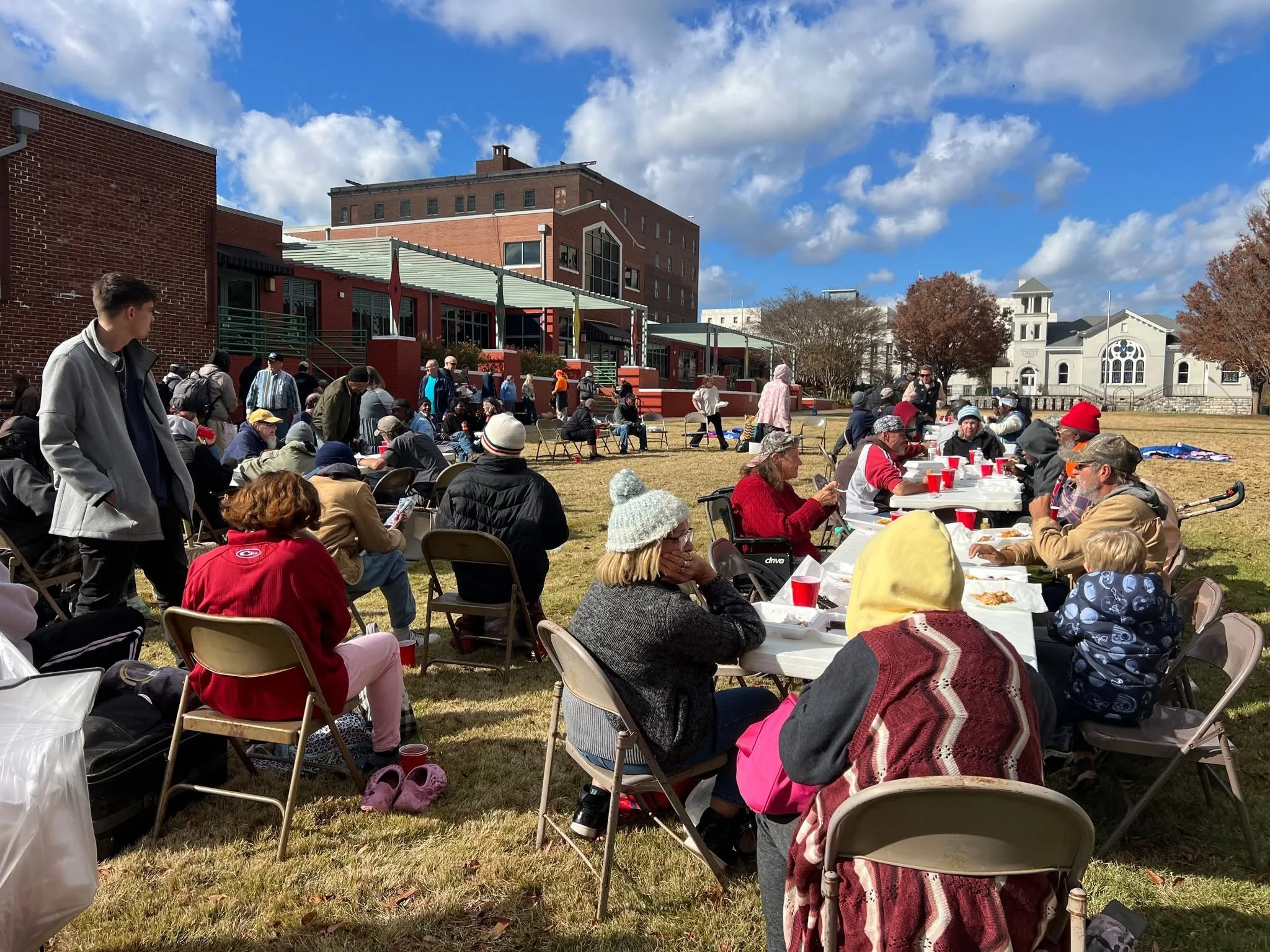 Outdoor gathering with people seated at tables with red cups, some standing, on a grassy area under a blue sky with clouds, near brick and white buildings.