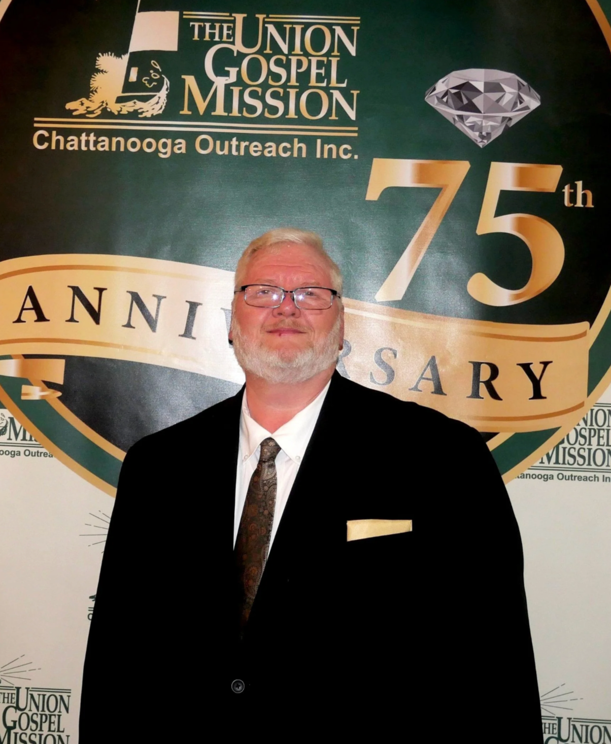 A man in a black suit and tie standing in front of a backdrop that reads 'The Union Gospel Mission Chattanooga Outreach Inc.' and celebrates its 75th anniversary, with a diamond graphic on top.