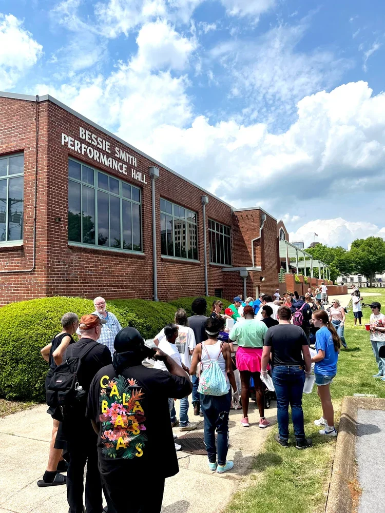 People standing in line outside the Bessie Smith Performance Hall building with a brick exterior and large windows, under a partly cloudy sky.